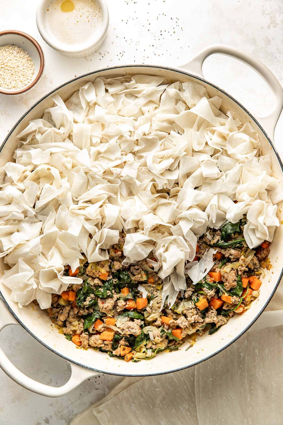 A round baking dish partly filled with cooked ground meat, carrots, spinach, and herbs, topped on one side with loosely crumpled phyllo dough sheets. Small bowls with oil and sesame seeds sit nearby.