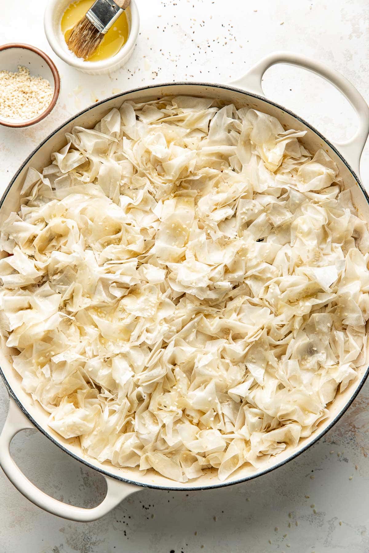 A round baking dish filled with crinkled, layered phyllo dough, lightly brushed with oil or butter. A small bowl of sesame seeds and a dish with a pastry brush and yellow liquid sit beside the dish.