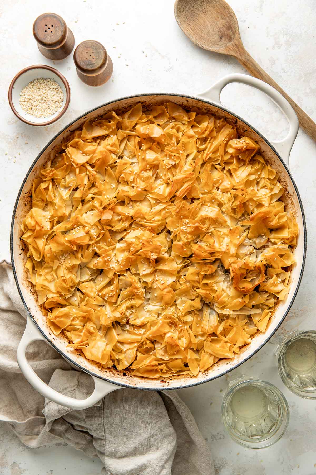 A round baking dish filled with golden, crispy phyllo pastry topped with sesame seeds sits on a light surface. Nearby are a wooden spoon, salt and pepper shakers, a small bowl of sesame seeds, and two glasses of white wine.
