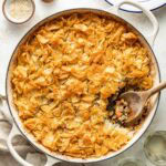 A round baking dish filled with a golden, crispy phyllo pastry pie, partially served to reveal a savory filling of meat and vegetables, with a wooden spoon inside. A plate, glasses, and small bowls are nearby.
