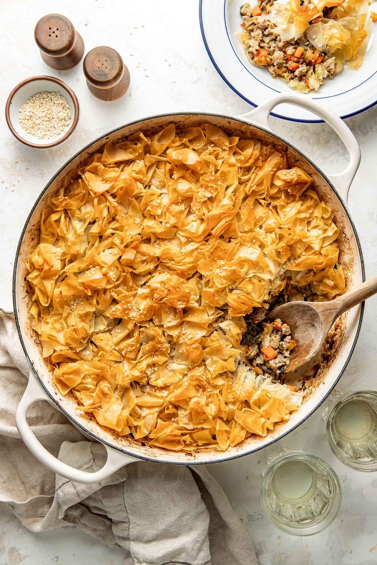 A round baking dish filled with a golden, crispy phyllo pastry pie, partially served to reveal a savory filling of meat and vegetables, with a wooden spoon inside. A plate, glasses, and small bowls are nearby.