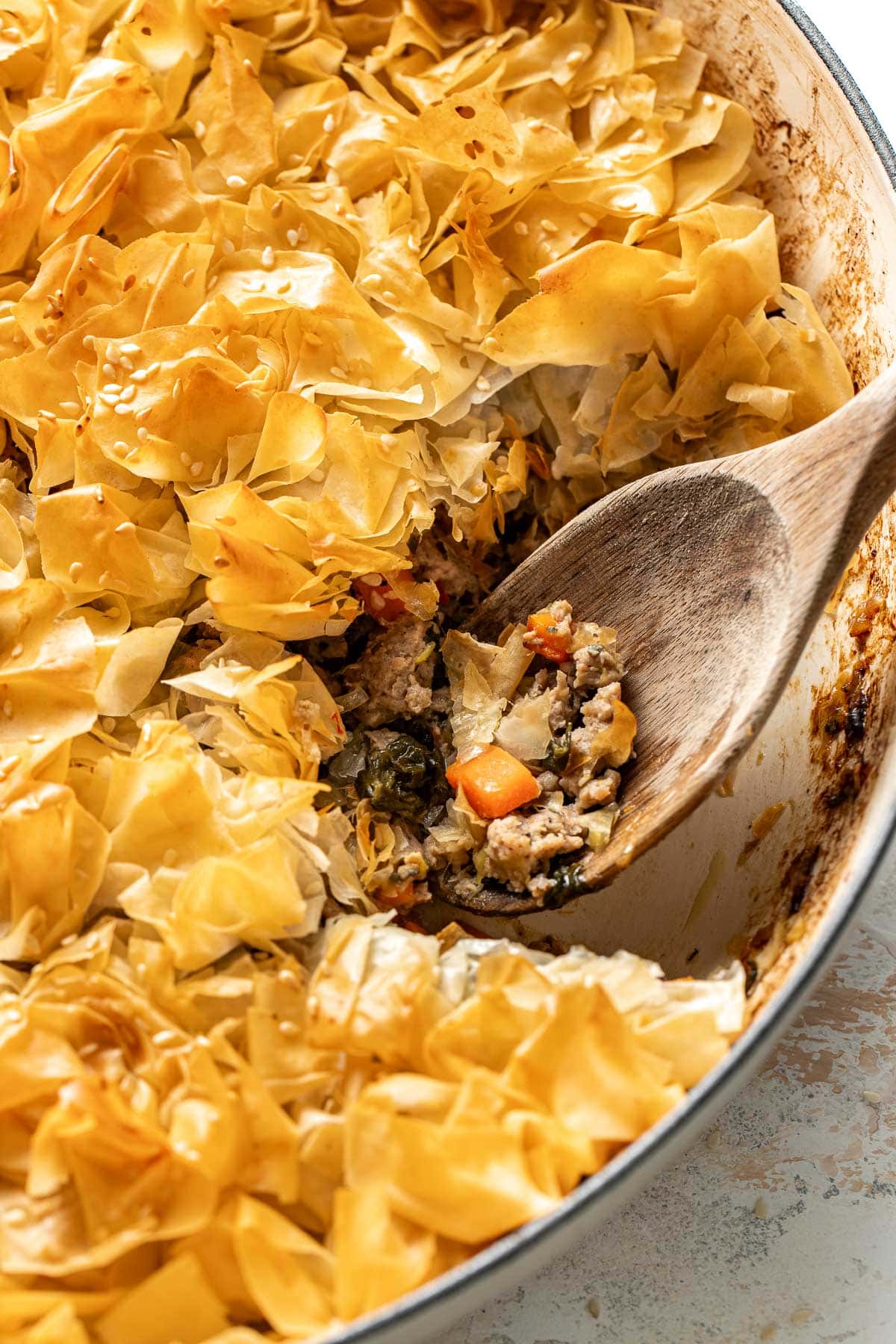 A close-up of a golden, flaky phyllo pastry pie in a round dish, with a wooden spoon scooping out a portion showing a savory vegetable filling with carrots and greens.