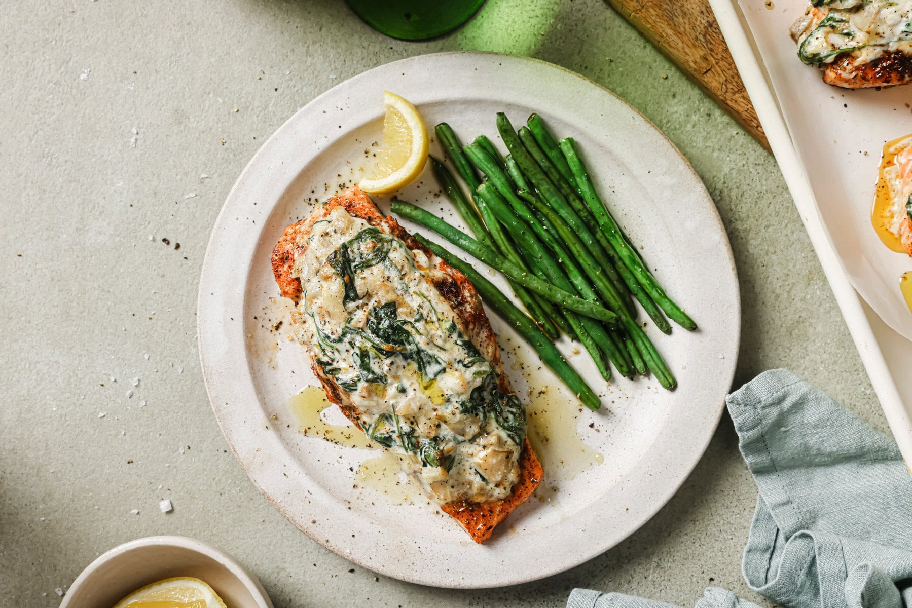 A white plate with a serving of baked salmon topped with creamy spinach sauce, a lemon wedge, and a side of green beans. A cloth napkin and extra lemon are nearby on a light, textured surface.