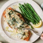 A plate with a piece of salmon topped with a creamy spinach sauce, served alongside steamed green beans and a lemon wedge. A hand is using a fork to cut into the salmon.