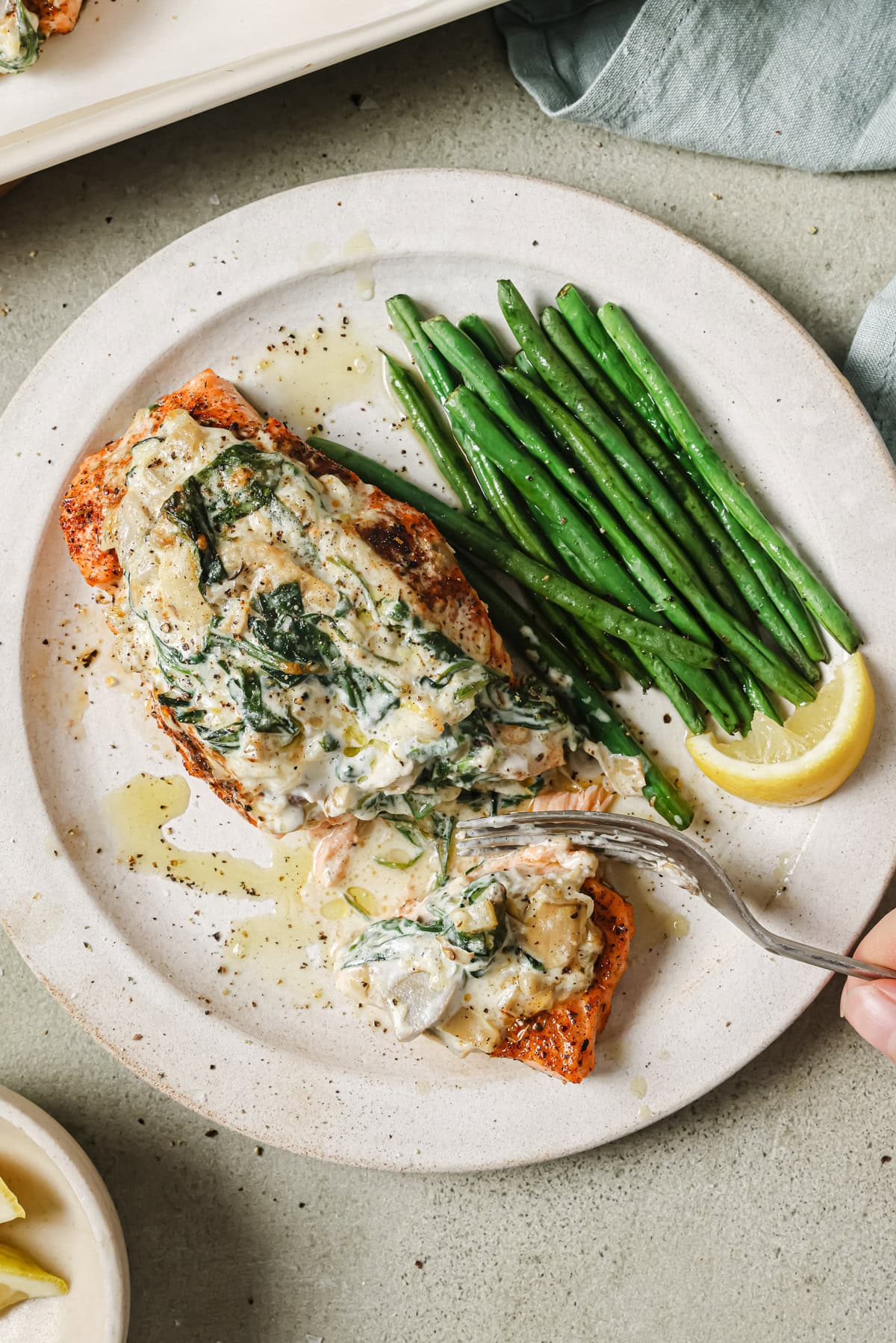 A plate with a piece of salmon topped with a creamy spinach sauce, served alongside steamed green beans and a lemon wedge. A hand is using a fork to cut into the salmon.