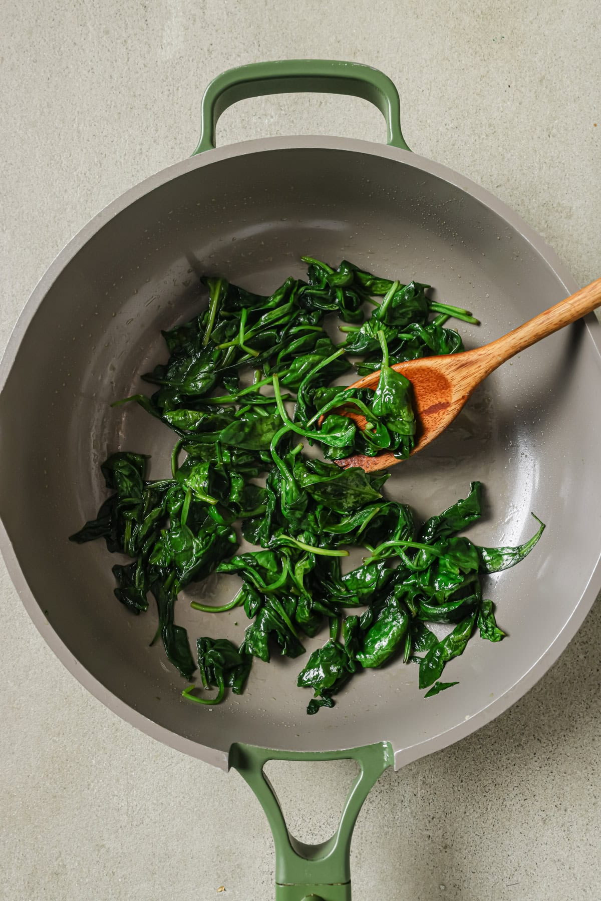 A sauté pan with cooked spinach being stirred by a wooden spoon, set on a light-colored surface.