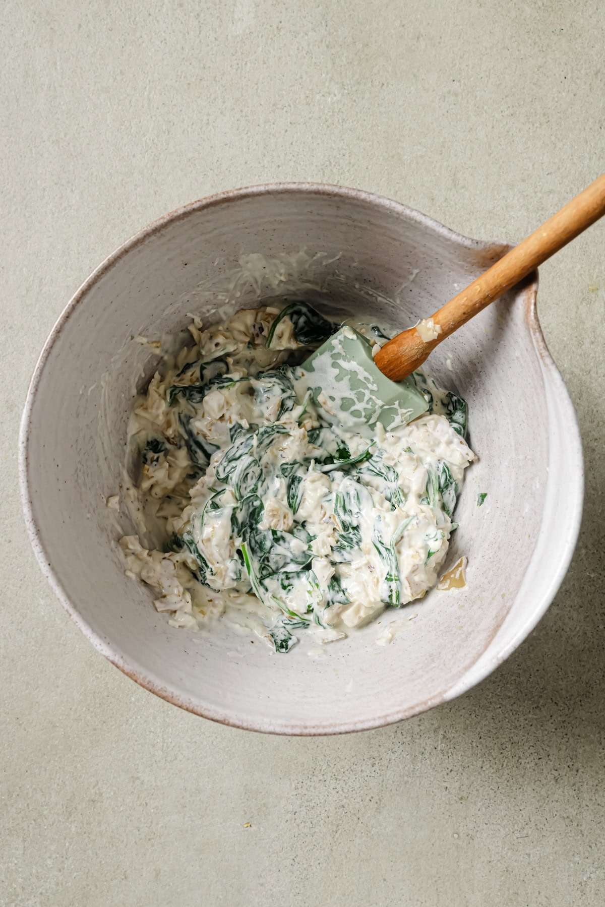 A mixing bowl containing a creamy spinach and cheese mixture being stirred with a green spatula on a light countertop.