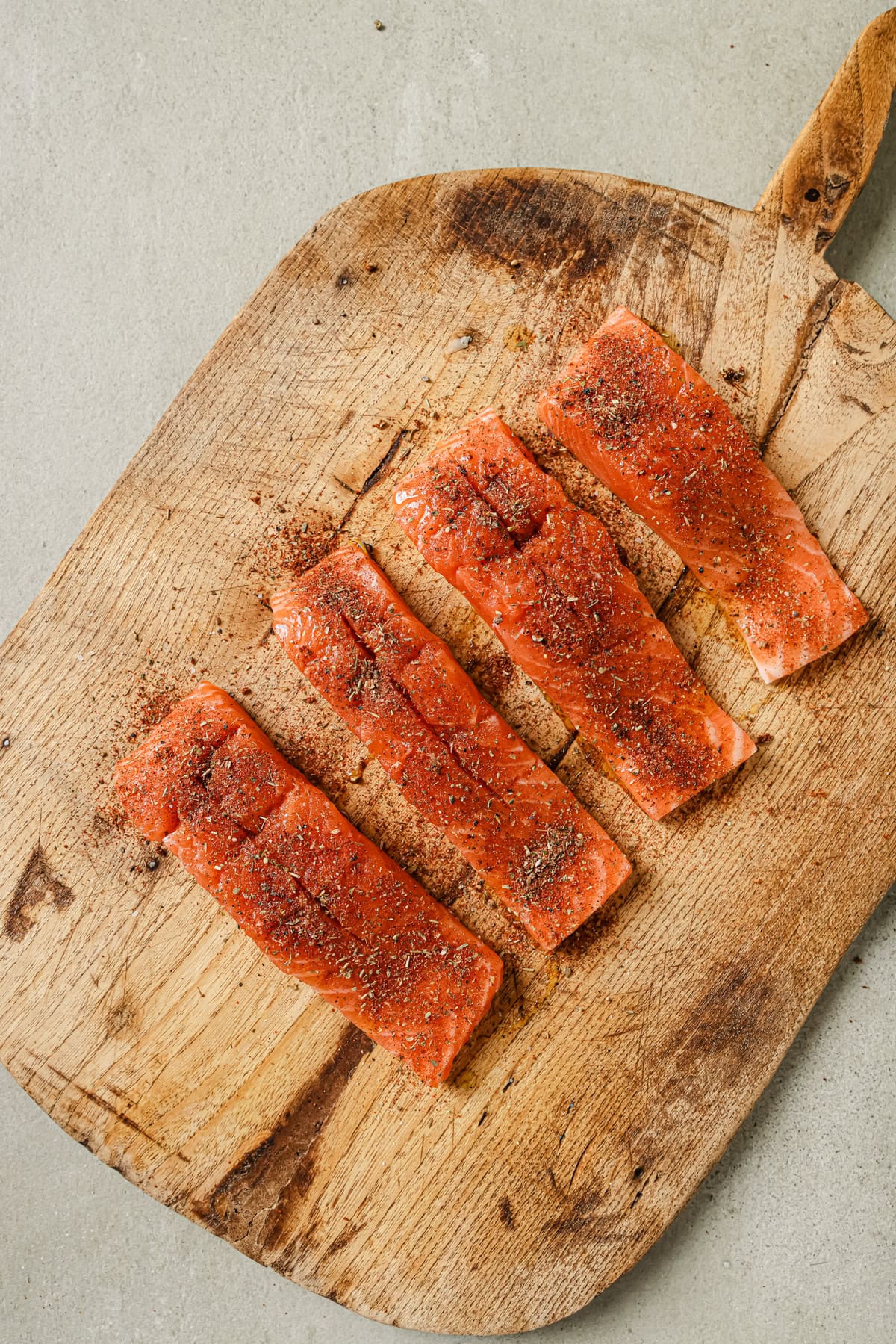 Four raw salmon fillets seasoned with herbs and spices are arranged on a wooden cutting board with a handle, placed on a light gray surface.