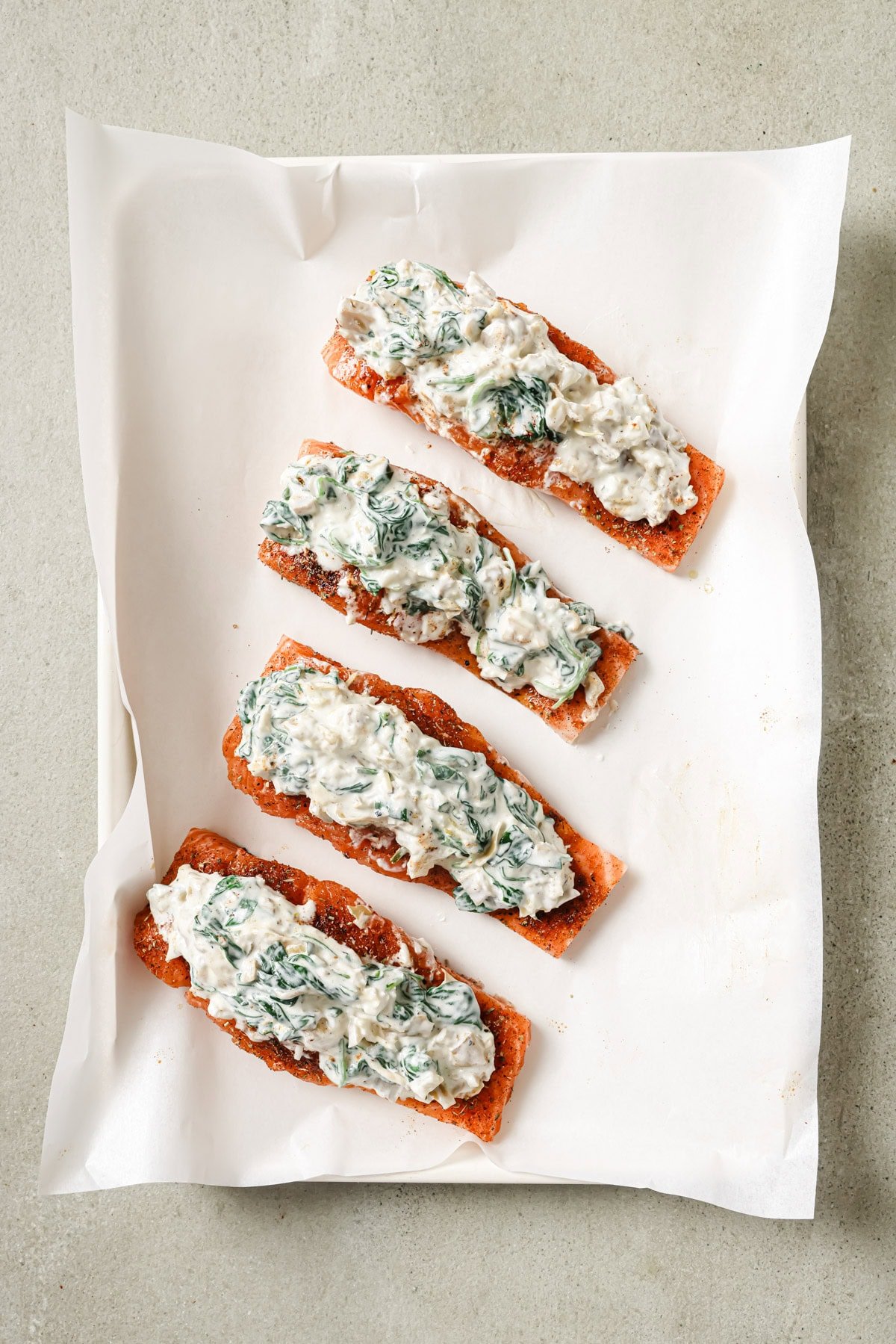 Four pieces of salmon fillet on parchment paper, each topped with a creamy spinach and cheese mixture, arranged in a row on a light-colored surface.