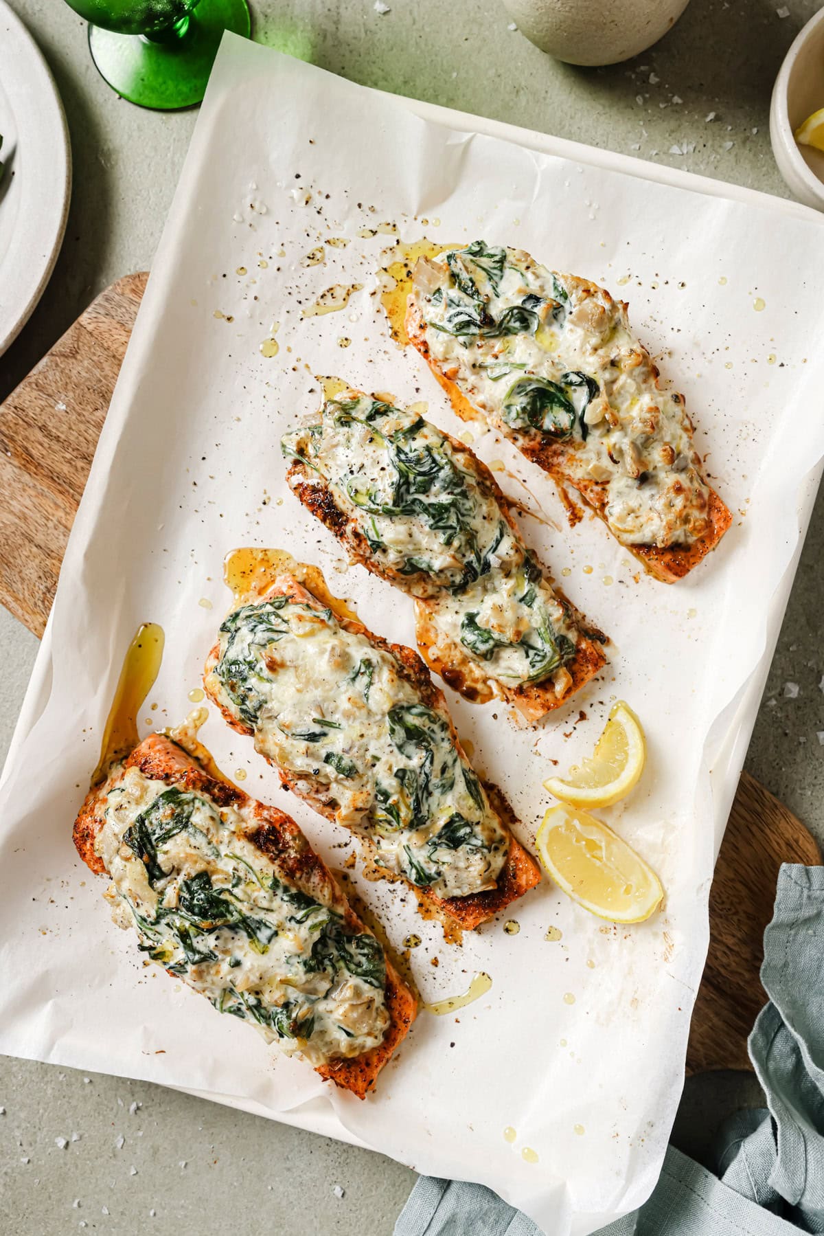 Four pieces of baked salmon topped with a creamy spinach mixture are arranged on parchment paper atop a wooden board, garnished with lemon wedges and a drizzle of oil.