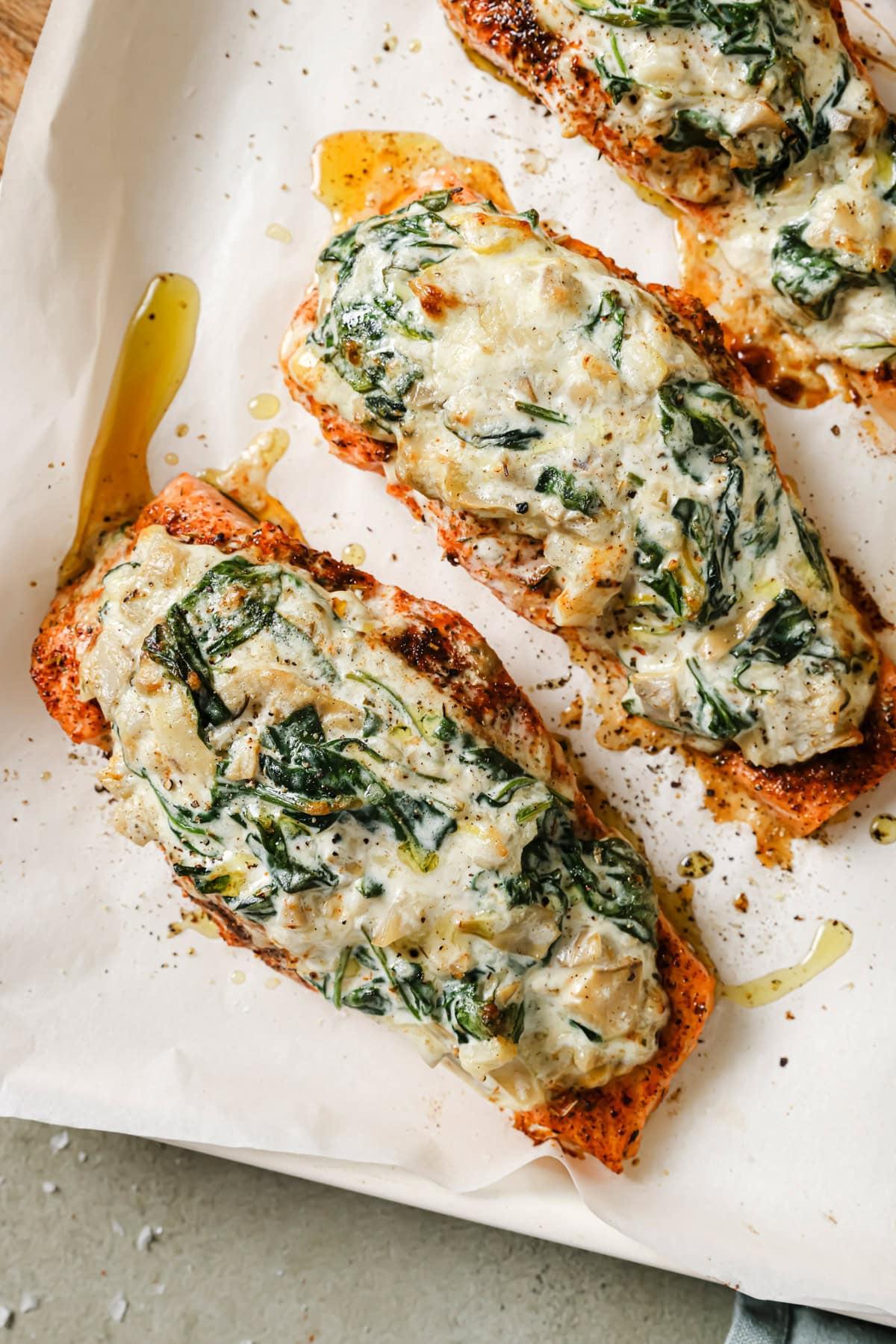 Three baked salmon fillets topped with a creamy spinach and cheese mixture, arranged on a parchment-lined baking sheet with a drizzle of oil and a sprinkle of black pepper.