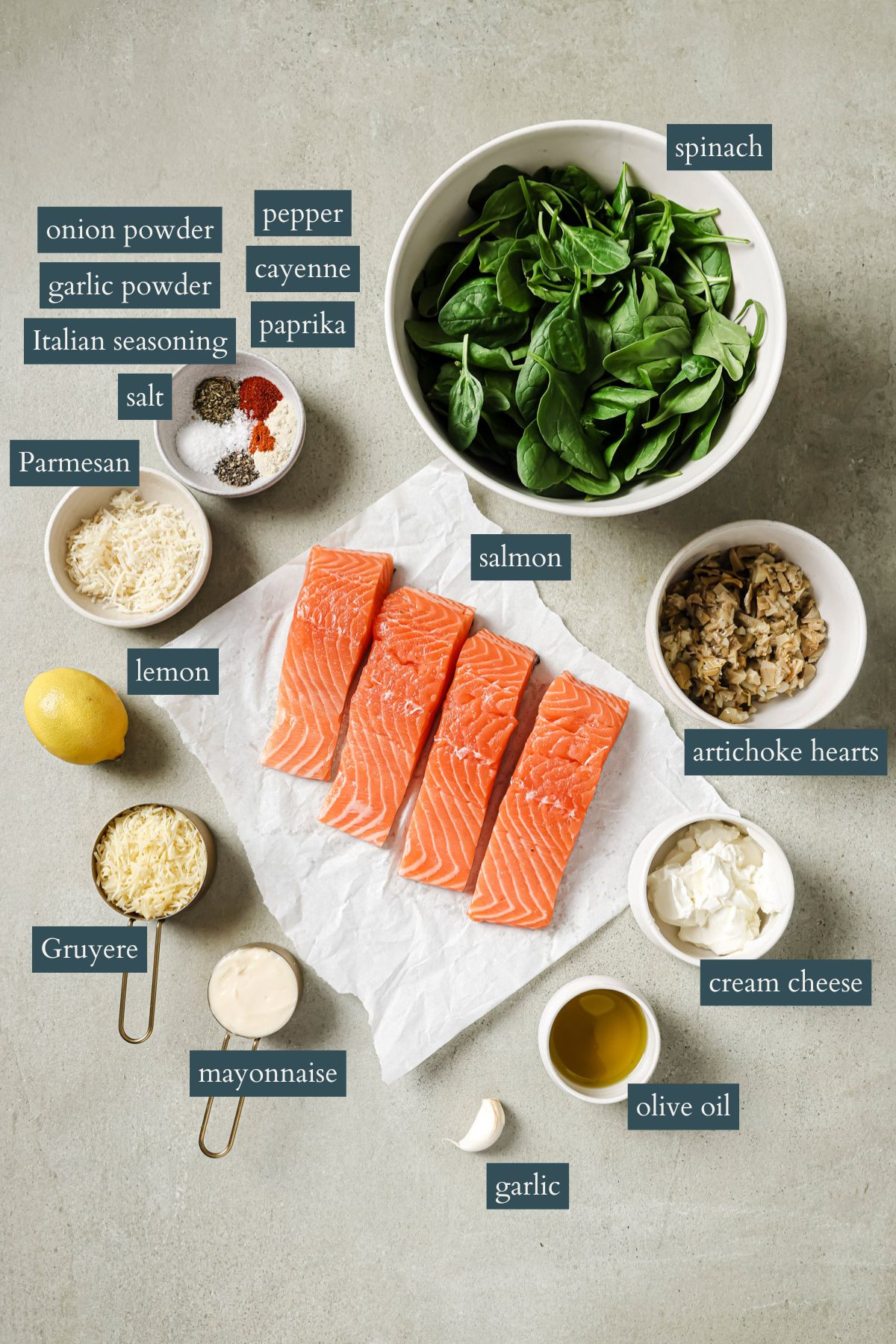 A flat lay of ingredients for a salmon recipe, including raw salmon fillets, spinach, artichoke hearts, Parmesan, Gruyere, garlic, lemon, mayonnaise, cream cheese, olive oil, and various seasonings in bowls.