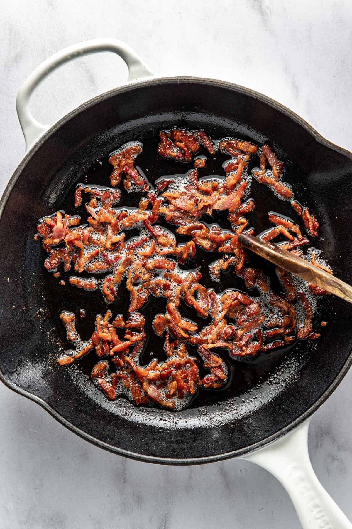 Crispy cooked bacon pieces in a black cast iron skillet with a white handle, being stirred by a wooden spoon on a light marble surface.