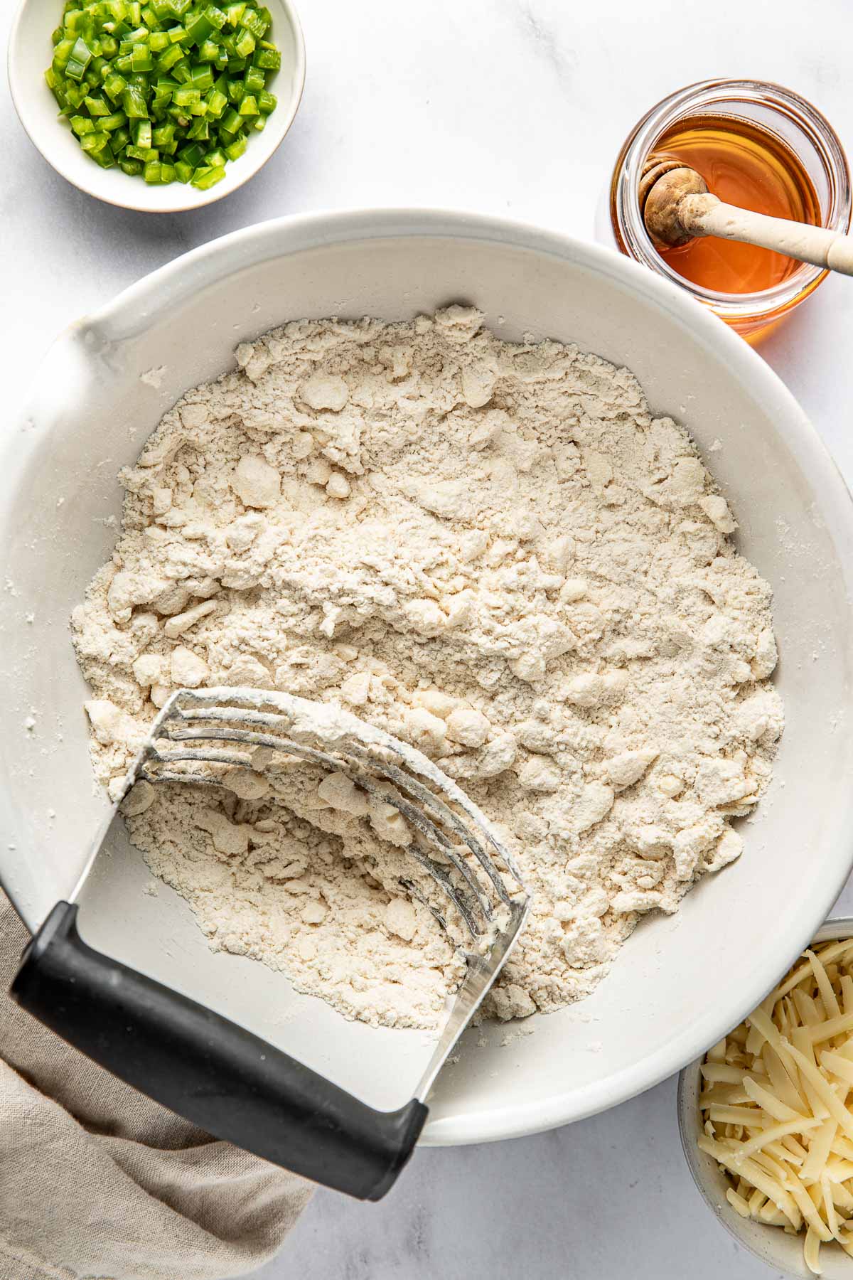 A bowl of flour mixture being mixed with a pastry cutter, surrounded by a small bowl of chopped green peppers, a jar of honey with a dipper, and a plate of shredded cheese on a white surface.