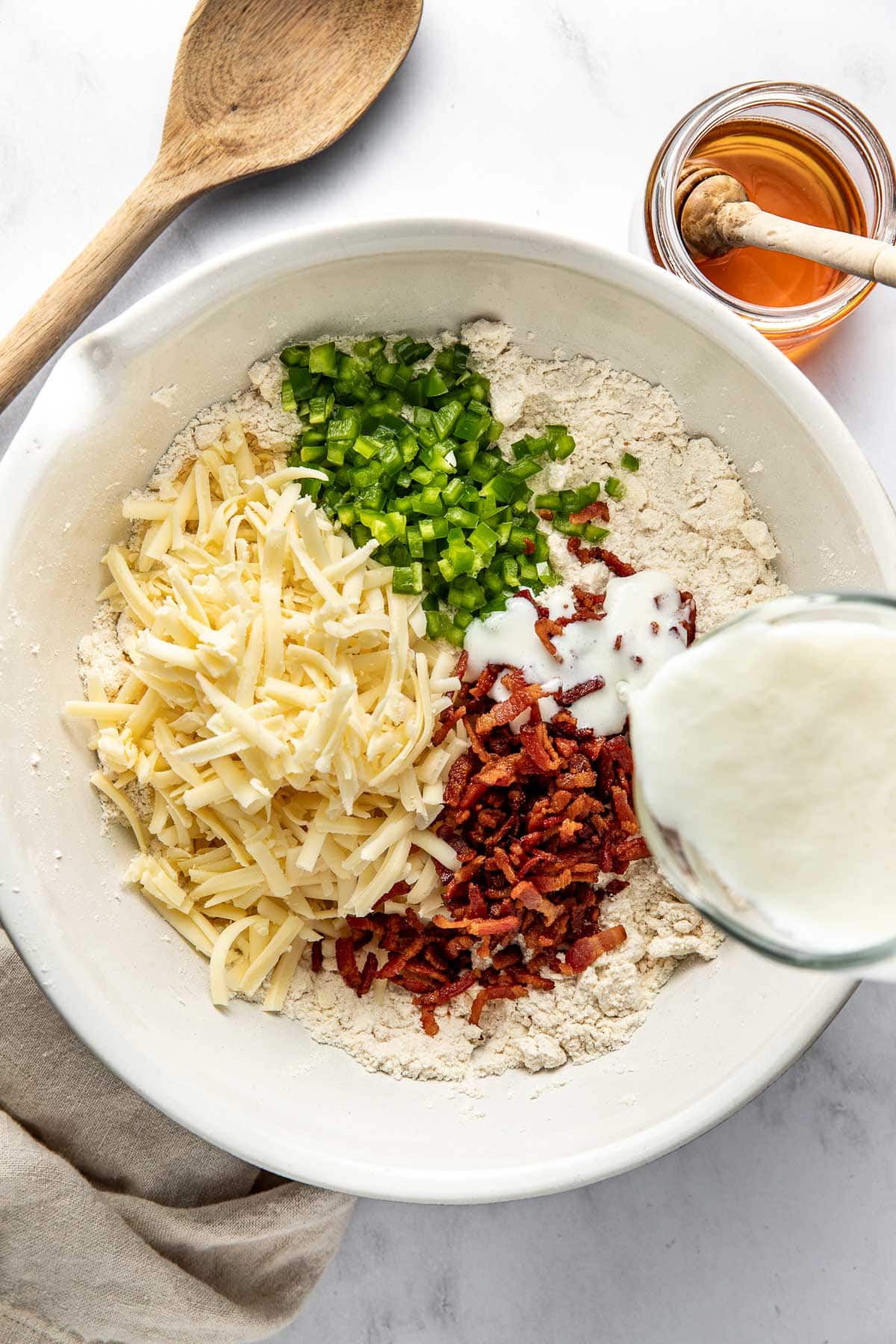 A bowl with flour, shredded cheese, chopped jalapeños, cooked bacon bits, and milk being poured in, next to a wooden spoon and a jar of honey on a white surface.