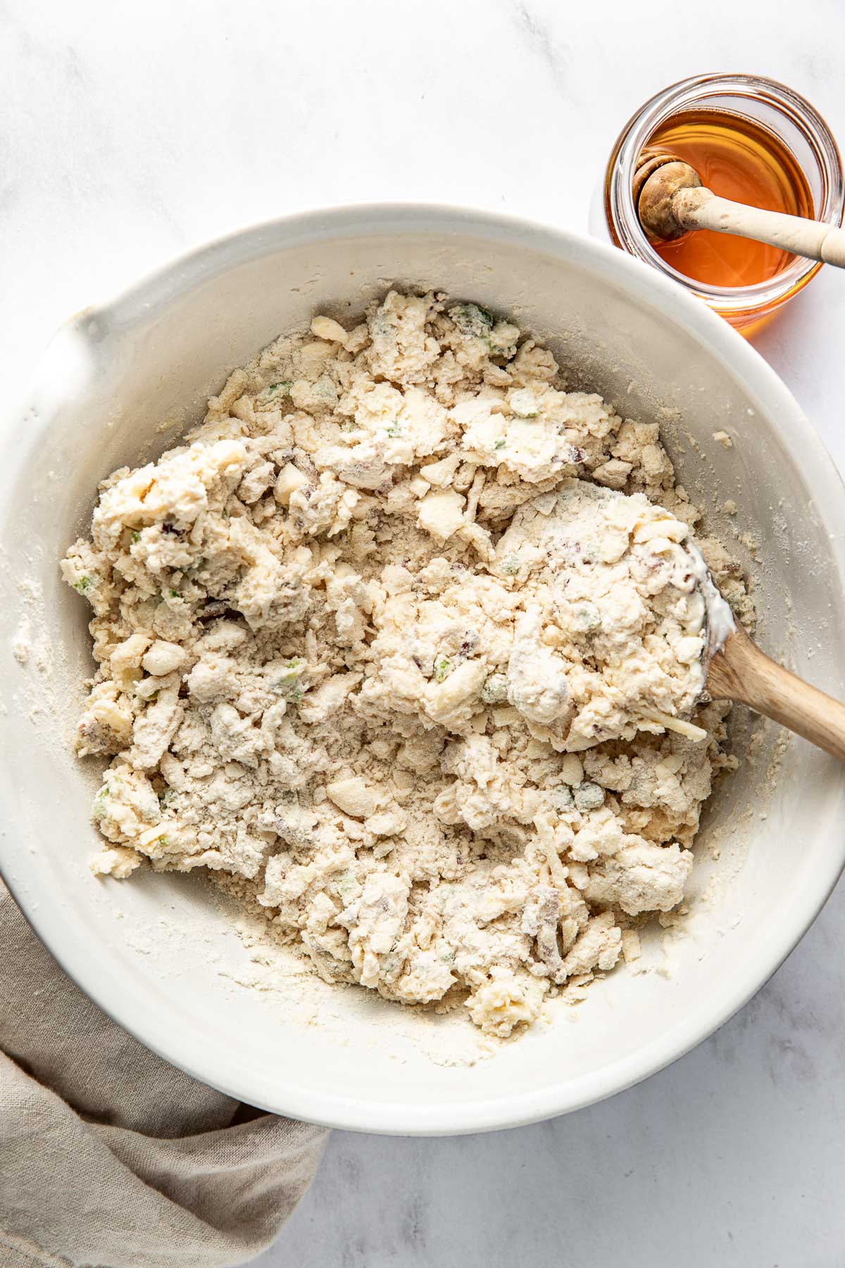A large white bowl filled with crumbly biscuit dough being mixed with a wooden spoon, next to a small jar of honey with a honey dipper on a white surface.