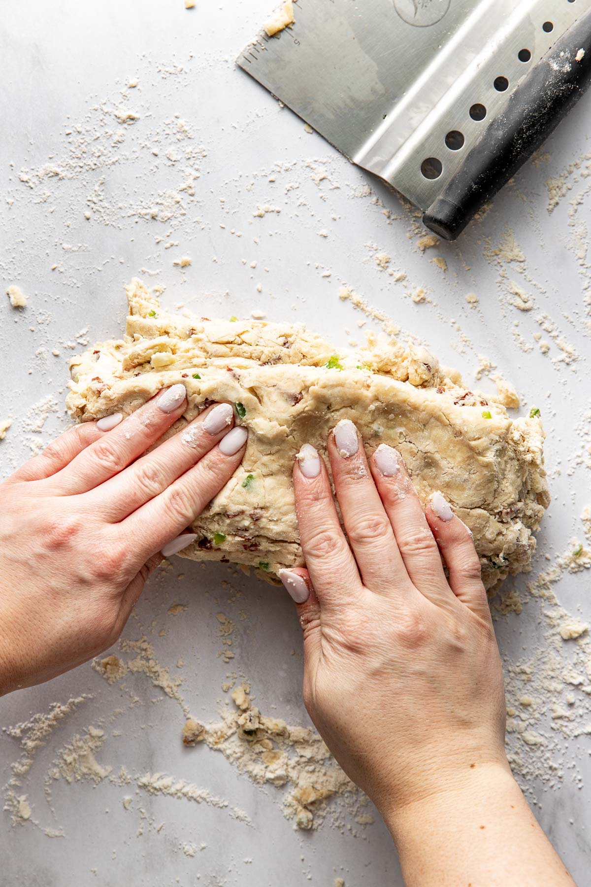 Two hands kneading dough with chopped green onions and bacon on a floured surface, next to a metal bench scraper.