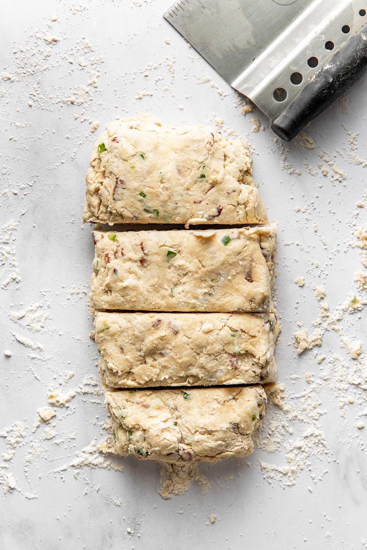 A block of biscuit dough with herbs and bacon is cut into four pieces on a floured surface. A metal bench scraper with a black handle rests beside the dough.