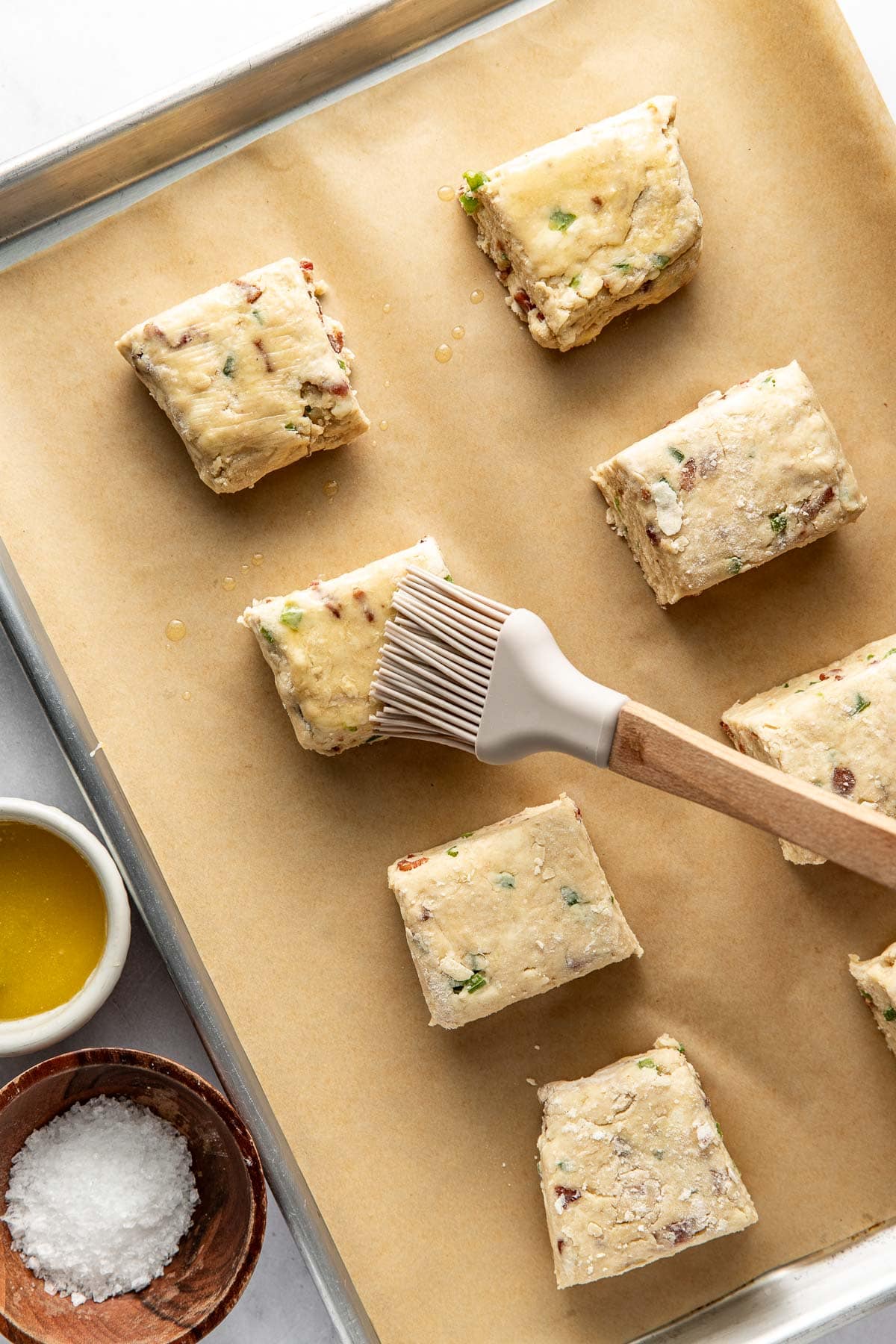 Unbaked scone dough squares on a parchment-lined baking sheet, being brushed with melted butter. Bowls of melted butter and coarse salt are nearby on a white surface.