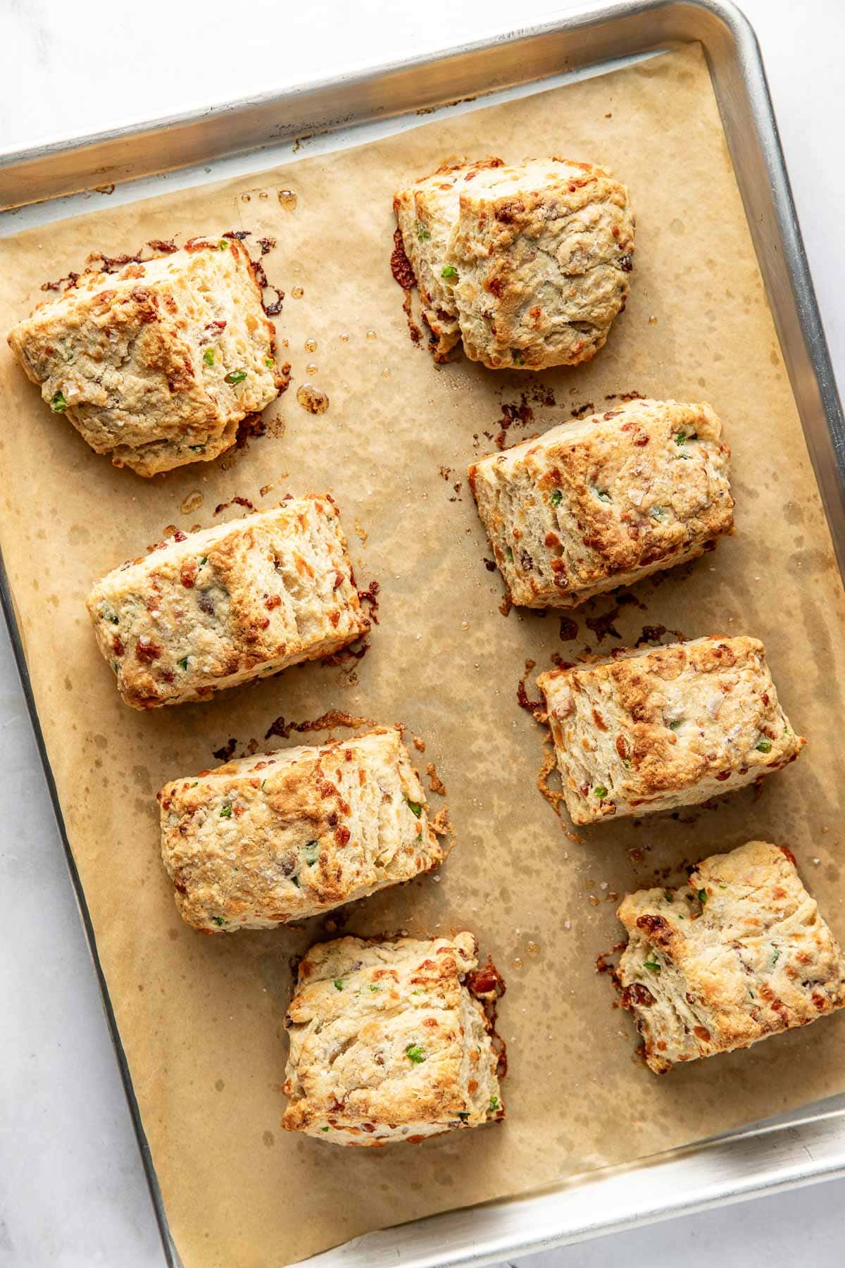 Eight golden-brown, square biscuits with visible bits of green and brown sit on a parchment-lined baking sheet. The biscuits look flaky with crisp edges and are spaced apart on the tray.