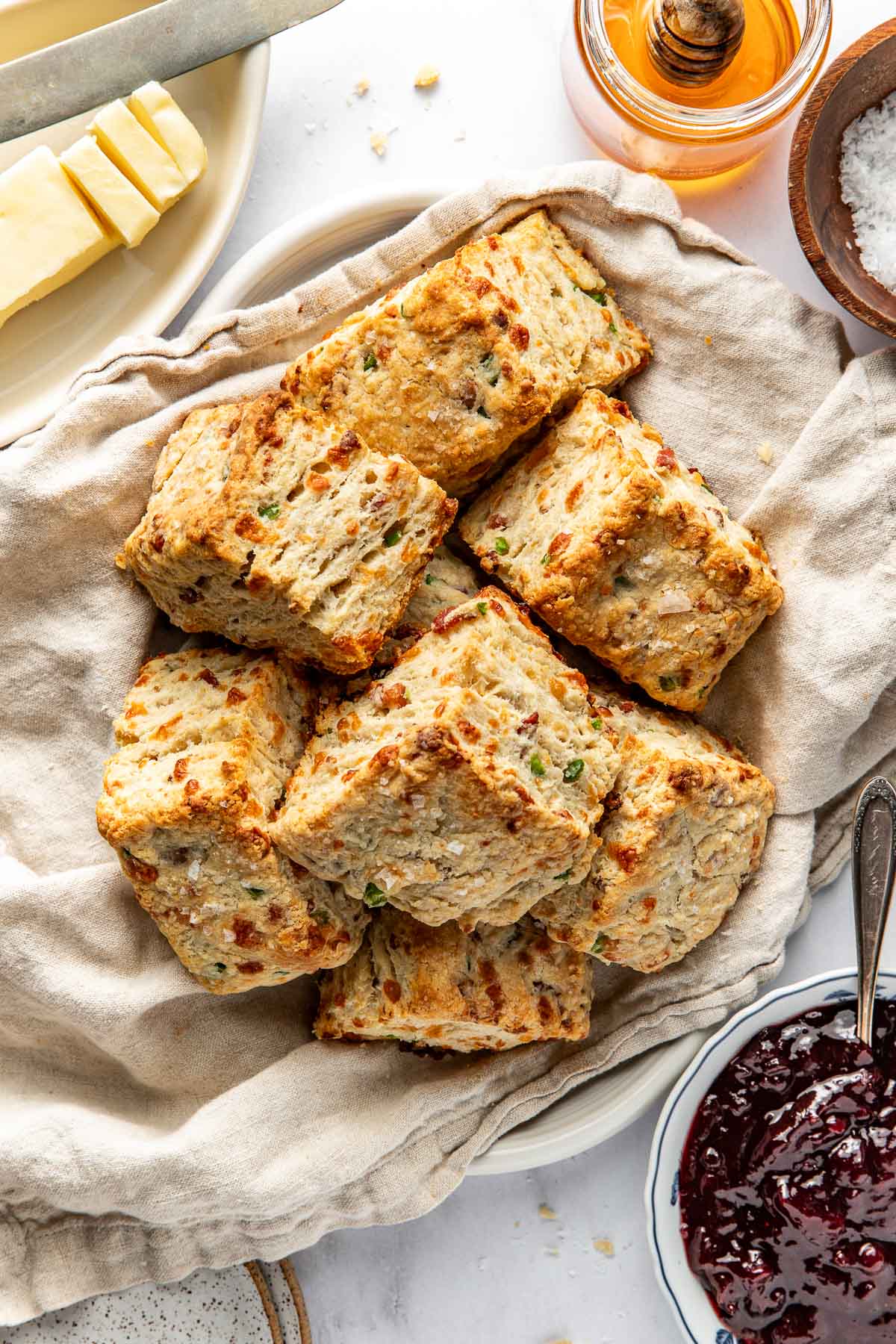 A basket lined with a beige cloth holds several golden-brown, flaky biscuits with visible green herbs. Surrounding the basket are plates with butter, honey, sea salt, and a bowl of dark red jam.