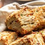 A close-up of freshly baked, flaky biscuits with visible bits of bacon and green herbs, stacked on a cloth. In the background, a plate with sliced butter is slightly out of focus.