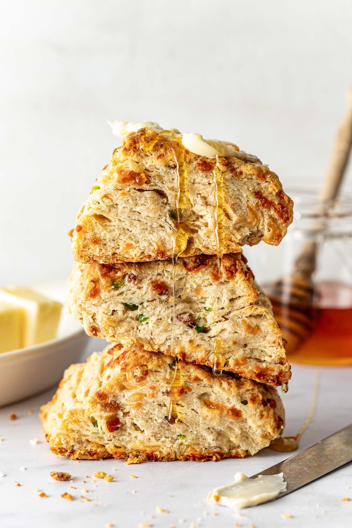 A stack of three cheddar and chive biscuits with crispy edges, drizzled with honey and topped with a pat of melting butter. A dish of butter and a jar of honey are in the background.