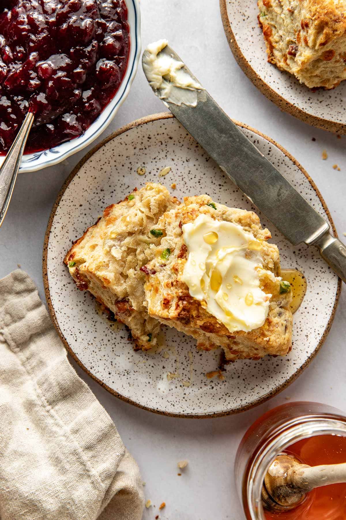 A plate with a split biscuit topped with butter and honey, a knife beside it, a jar of honey with a dipper, a bowl of berry jam, and a cloth napkin on a light surface.
