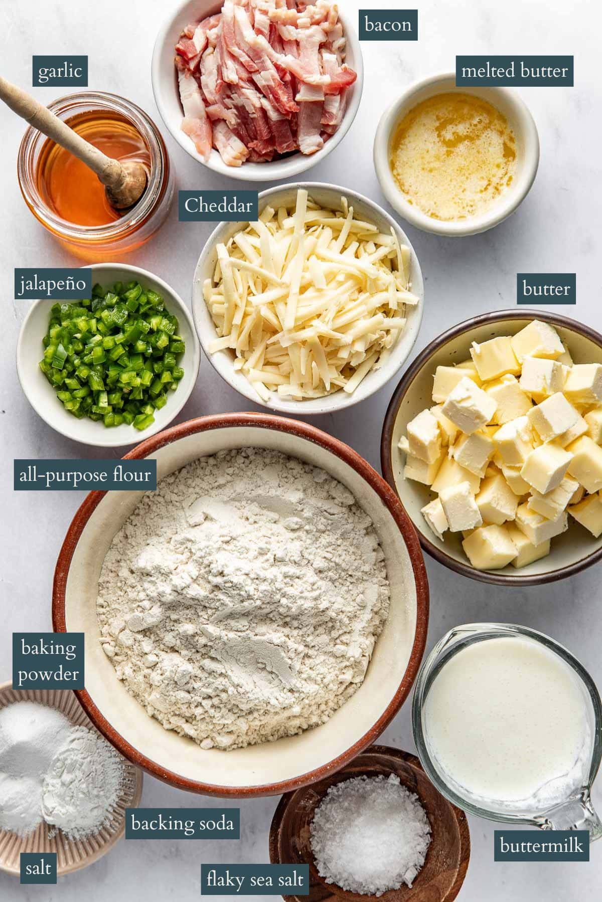 Overhead view of labeled ingredients in bowls: chopped garlic, cooked bacon, melted butter, shredded cheddar, chopped jalapeño, cubed butter, all-purpose flour, baking powder, baking soda, flaky sea salt, and buttermilk.
