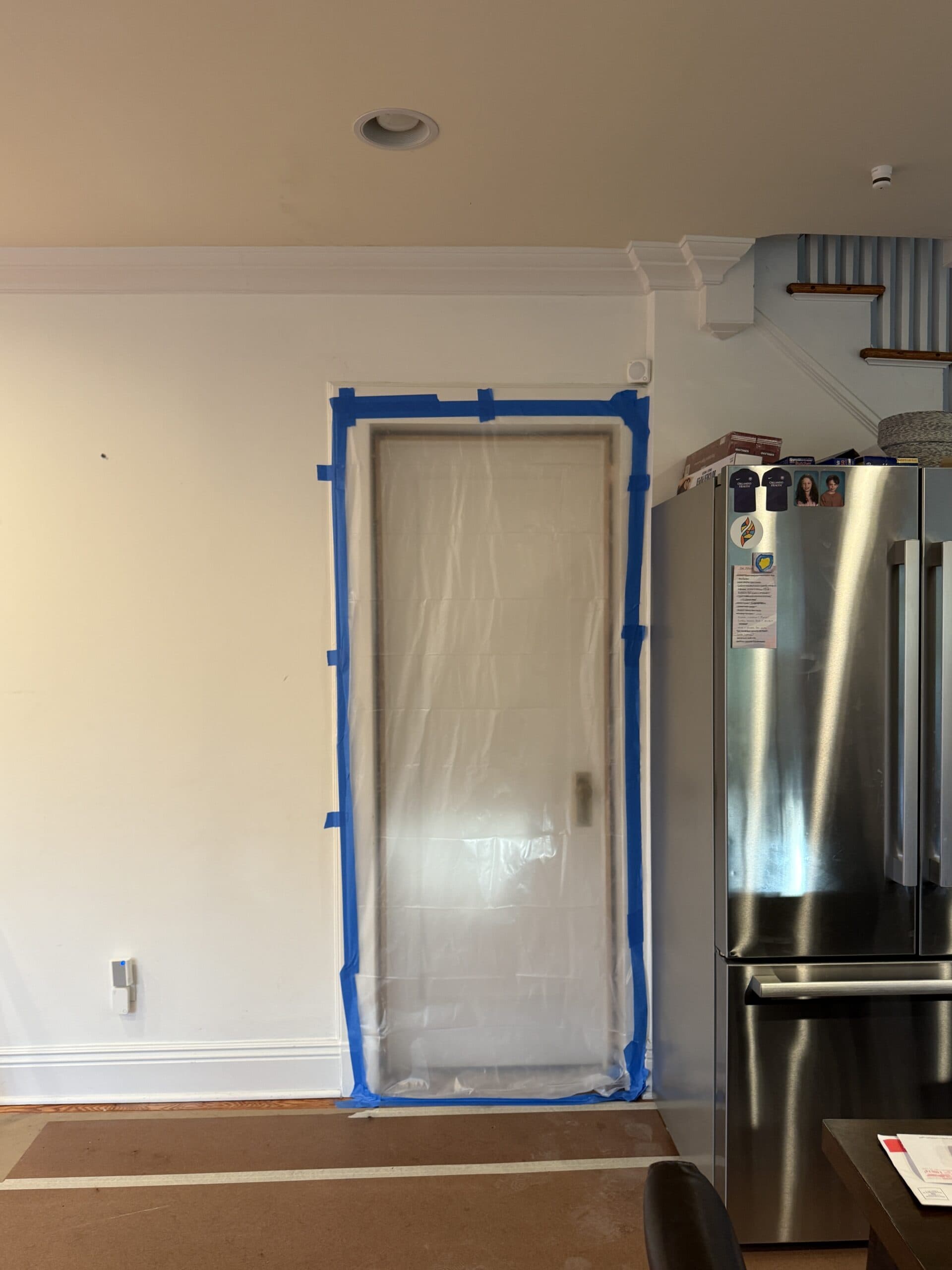 A doorway is covered with clear plastic sheeting secured by blue painter’s tape. The area is next to a stainless steel refrigerator and a staircase, in a partially renovated room.