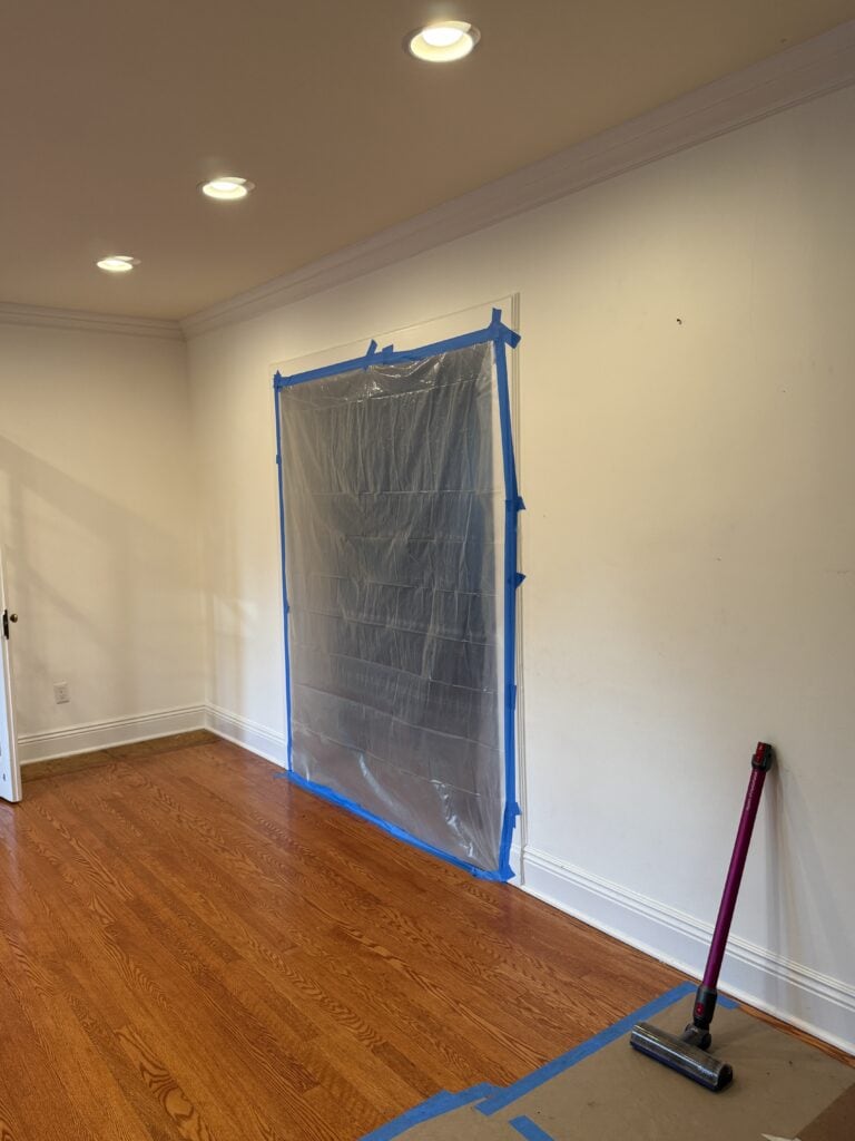 A room with hardwood floors and white walls, featuring a large plastic sheet taped over a doorway, possibly for dust containment during renovation. Ceiling lights are on, and a vacuum cleaner stands in the corner.
