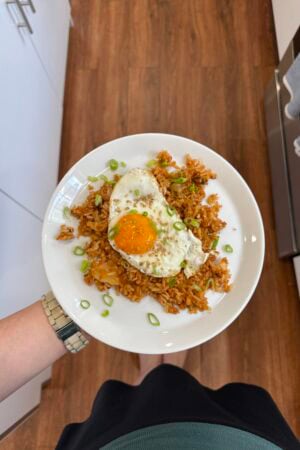 A person holding a white plate with fried rice topped with a sunny-side-up egg and chopped green onions, standing on a wooden floor in a kitchen.