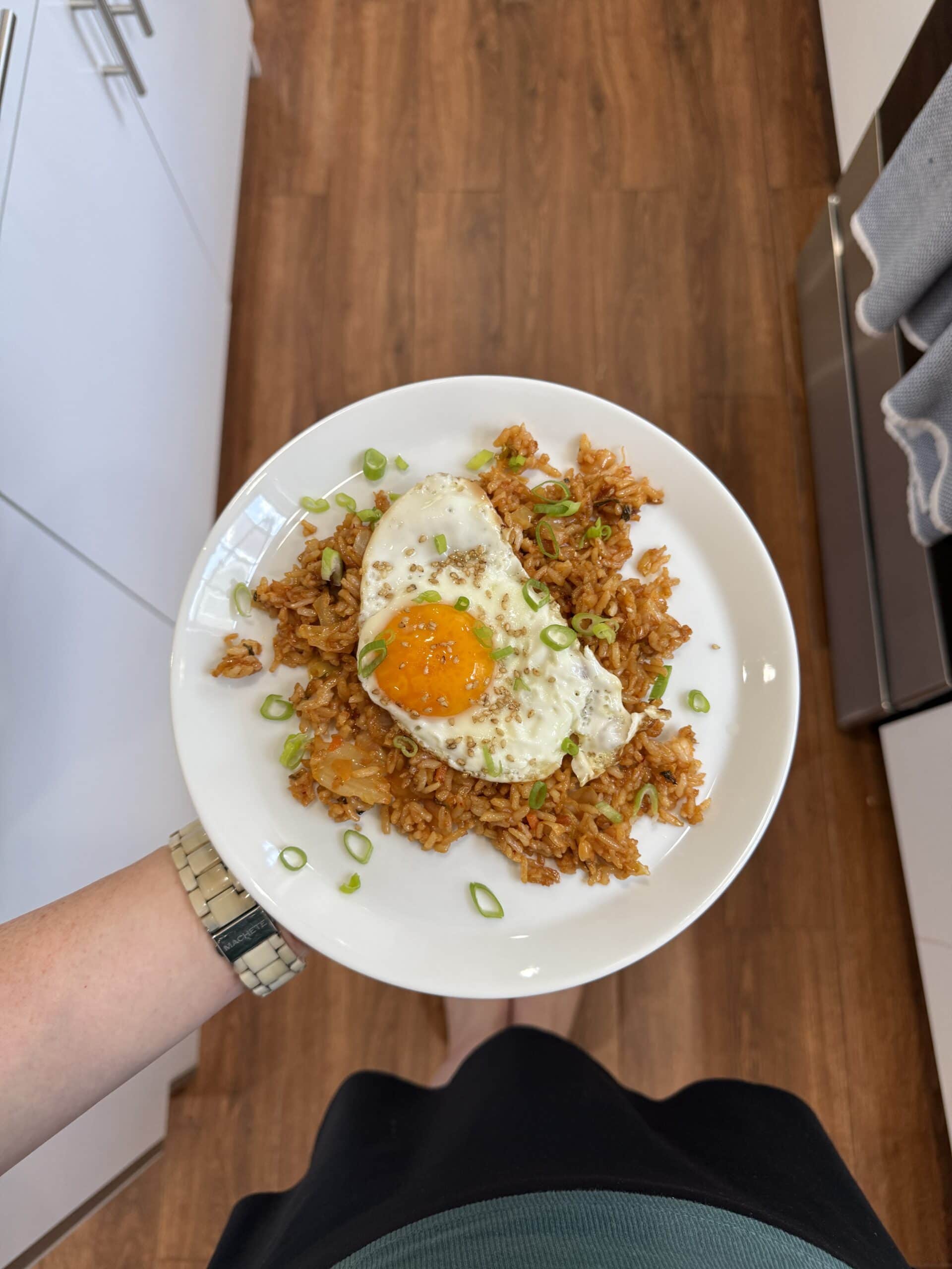 A person holding a white plate with fried rice topped with a sunny-side-up egg and chopped green onions, standing on a wooden floor in a kitchen.