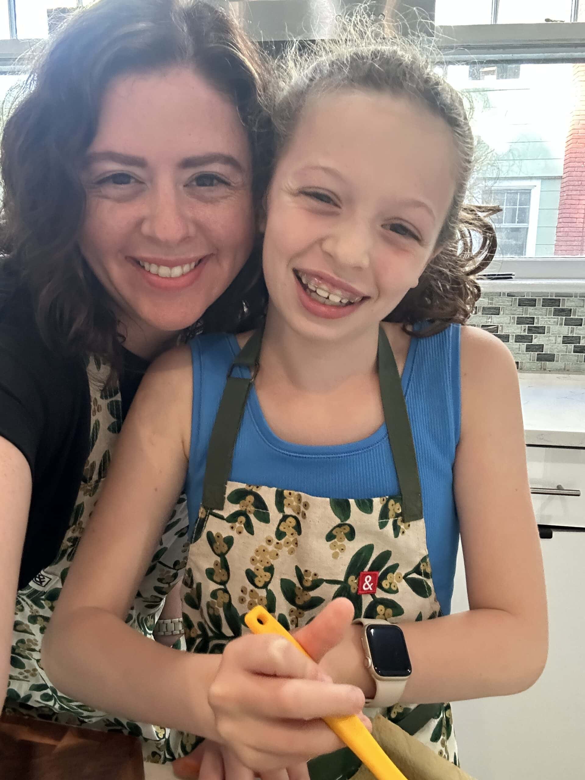 Two people, an adult and a child, smile at the camera in a bright kitchen while wearing matching aprons. The child holds a yellow spatula and wears a smartwatch. Sunlight streams through a window behind them.
