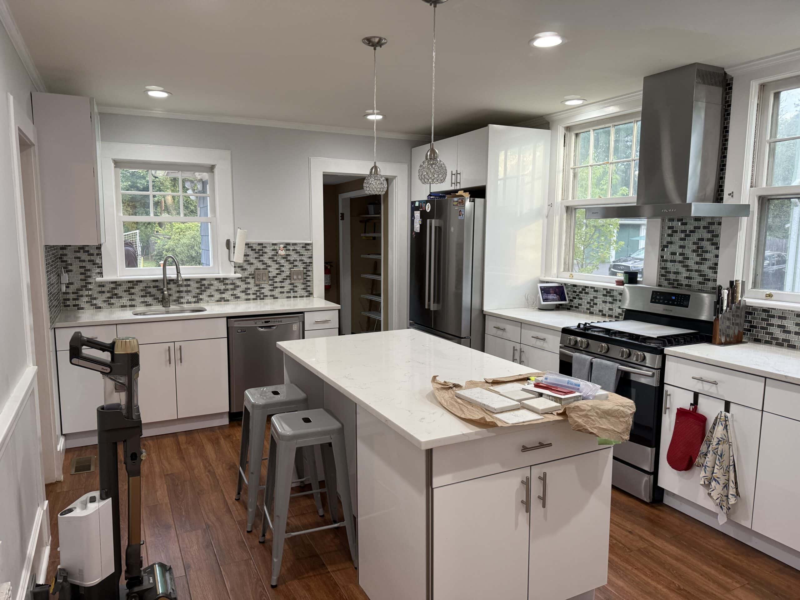 Bright modern kitchen with white cabinets, marble island, stainless steel appliances, two gray bar stools, a vacuum cleaner, and a tiled backsplash. Sunlight streams through large windows above the sink and stove.
