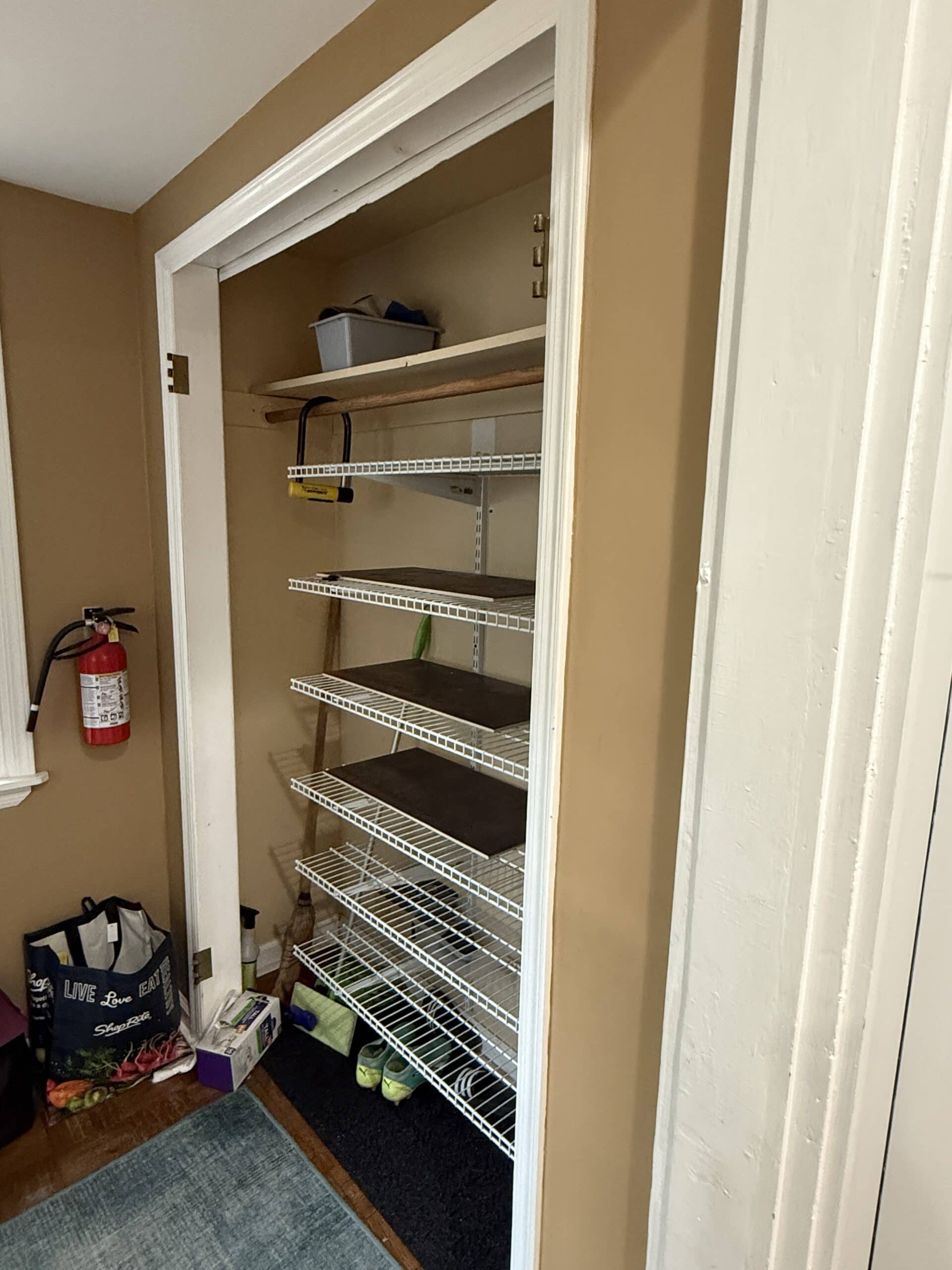 A closet with tan walls and white trim contains white wire shelves holding a few items, including cleaning supplies and shoes. A fire extinguisher hangs nearby, and bags sit on the floor outside the closet.