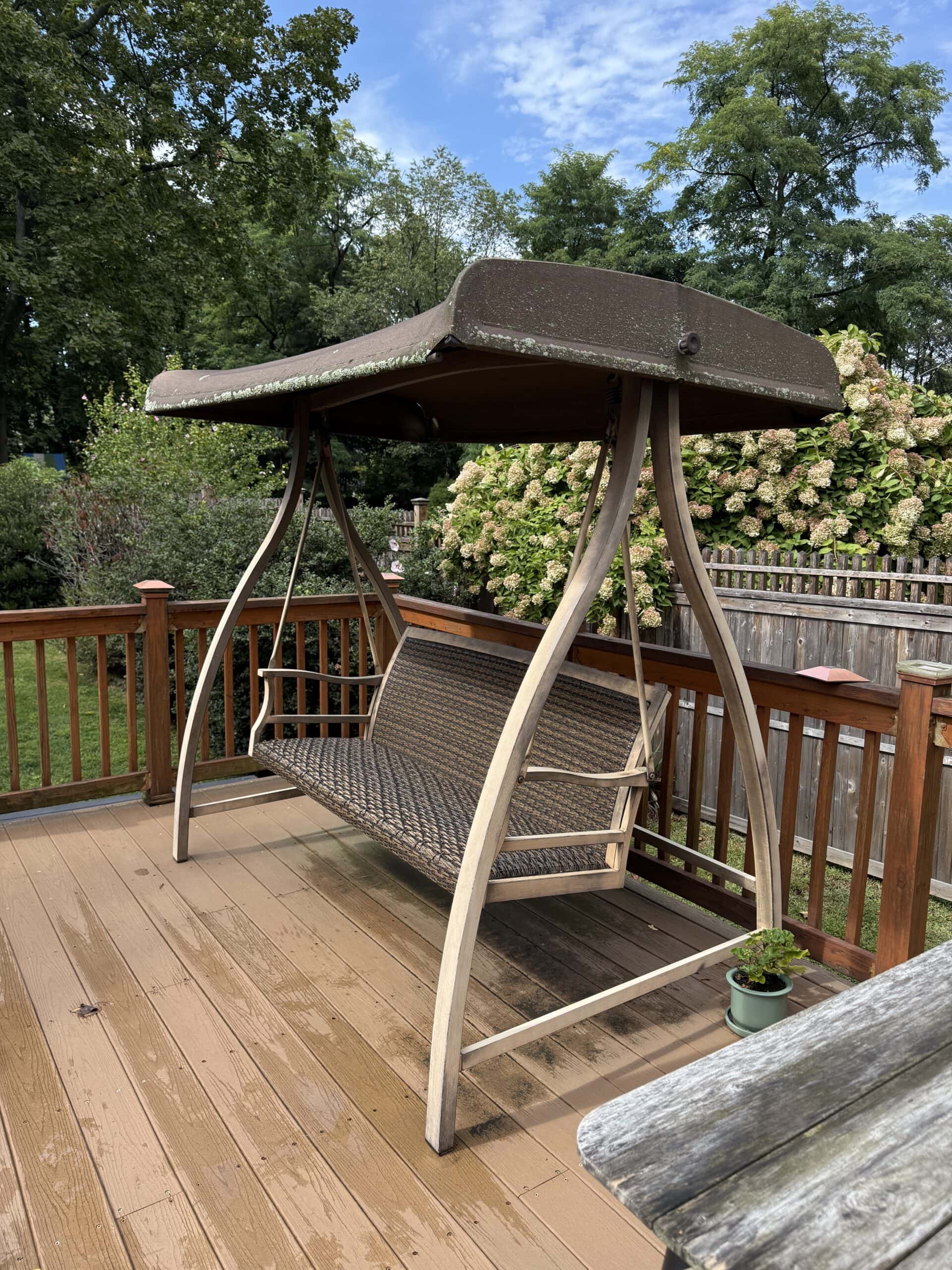 A brown outdoor swing with a canopy sits on a wooden deck surrounded by a wooden railing. There are green trees and bushes in the background, and a small potted plant is on the deck.
