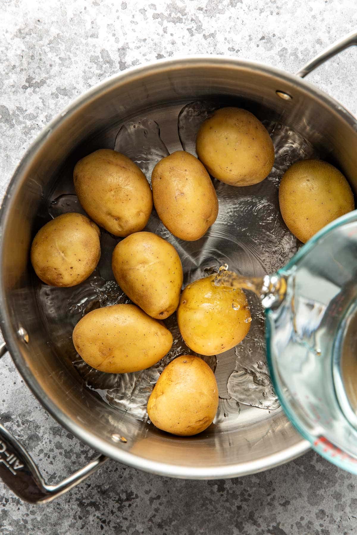 A stainless steel pot contains several whole, unpeeled potatoes. Water is being poured into the pot from a glass measuring cup. The surface below is gray and textured.