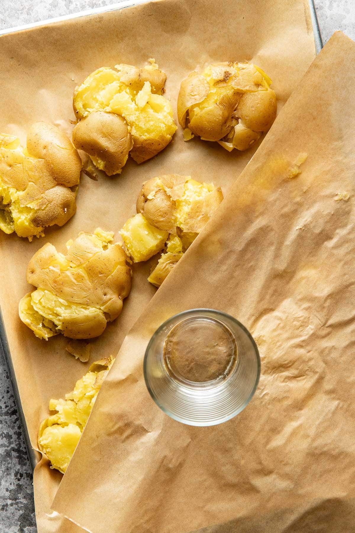 A baking tray lined with parchment paper holds several smashed potatoes. A clear drinking glass, used for smashing, is resting on the tray. Another sheet of parchment paper is partially covering the potatoes.