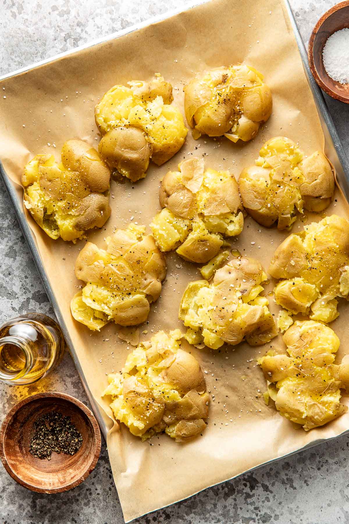A baking tray lined with parchment paper holds several smashed, roasted potatoes seasoned with black pepper. Nearby are small bowls with salt, pepper, and a bottle of olive oil.