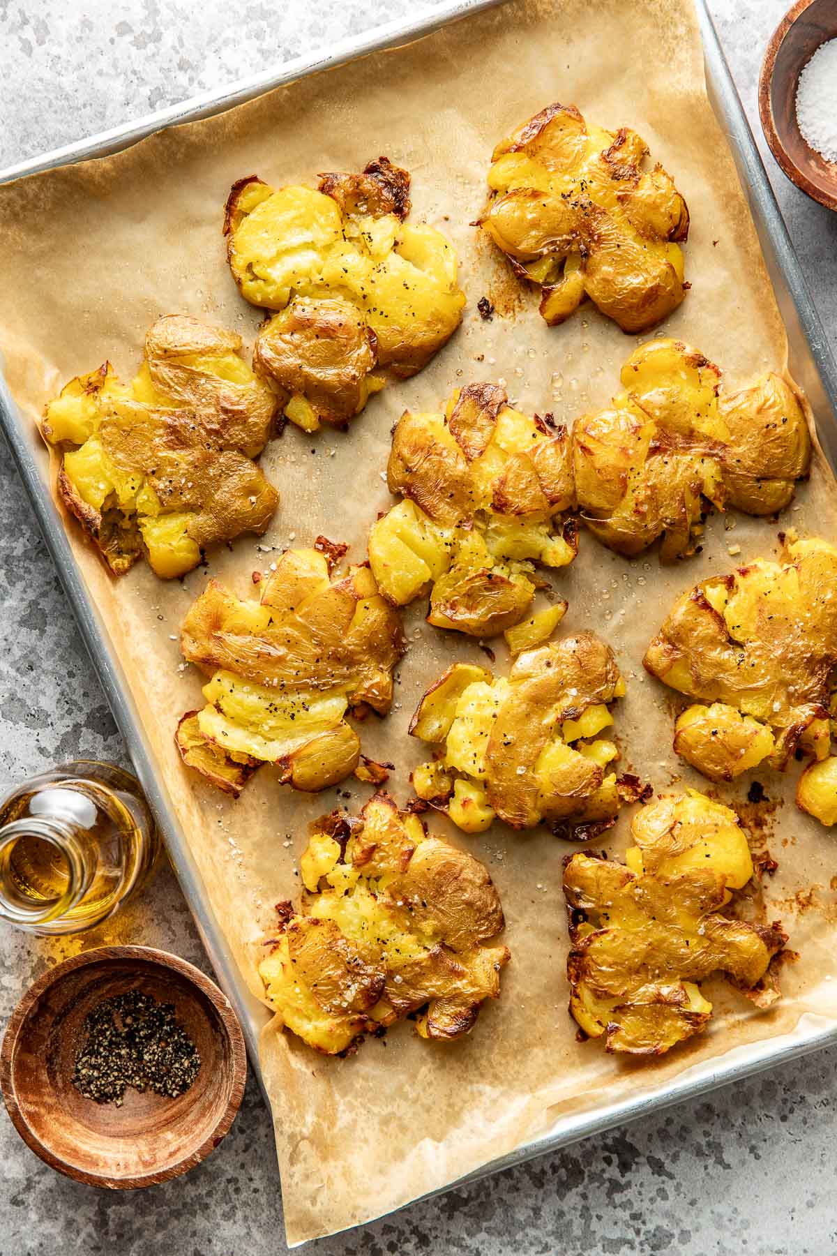 A baking sheet lined with parchment paper holds several smashed, roasted potatoes seasoned with salt and pepper. Nearby are small bowls of salt, pepper, and a small bottle of olive oil on a gray countertop.