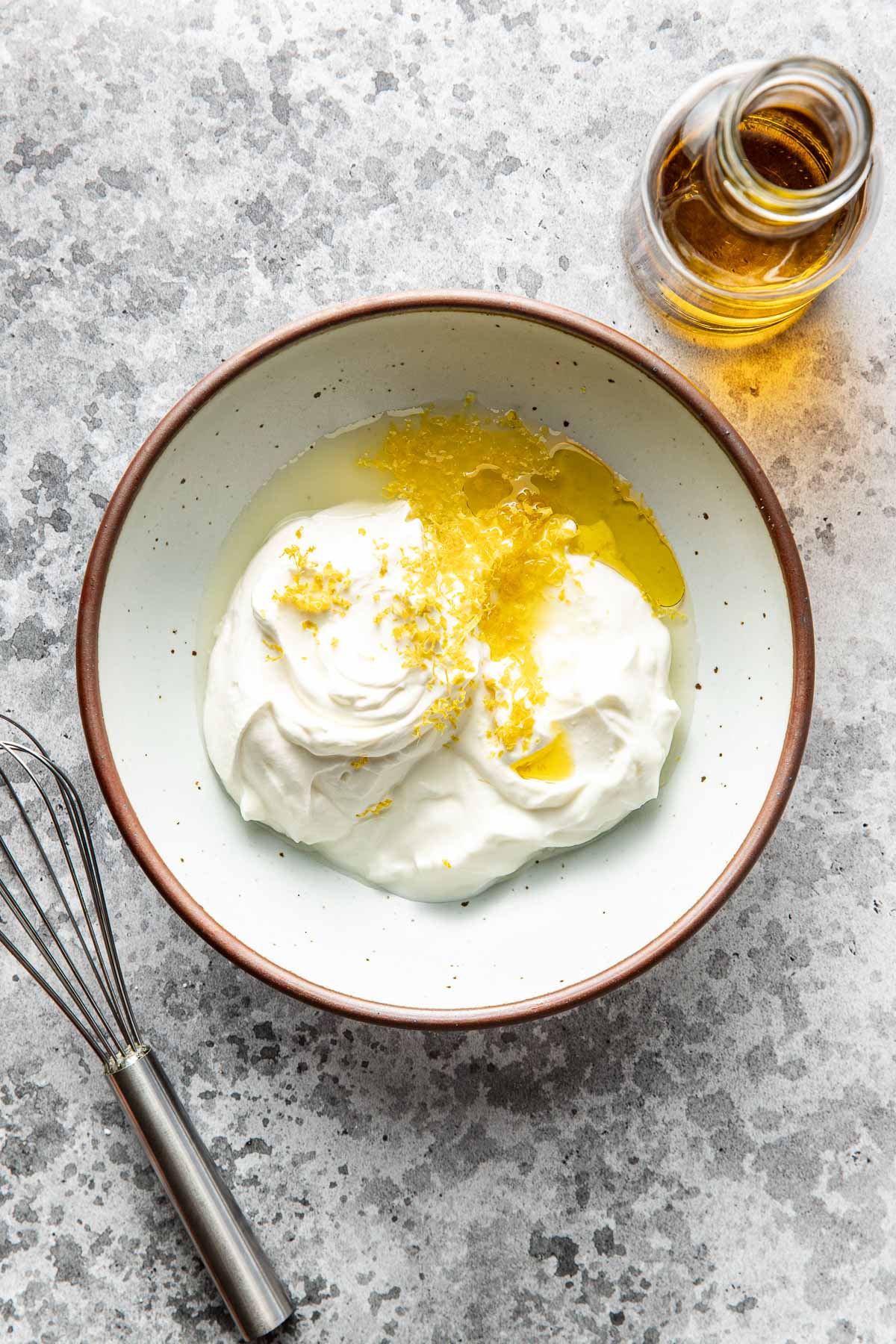 A bowl of whipped cream topped with lemon zest and honey sits on a gray textured surface. A small glass jar of honey and a metal whisk are nearby.