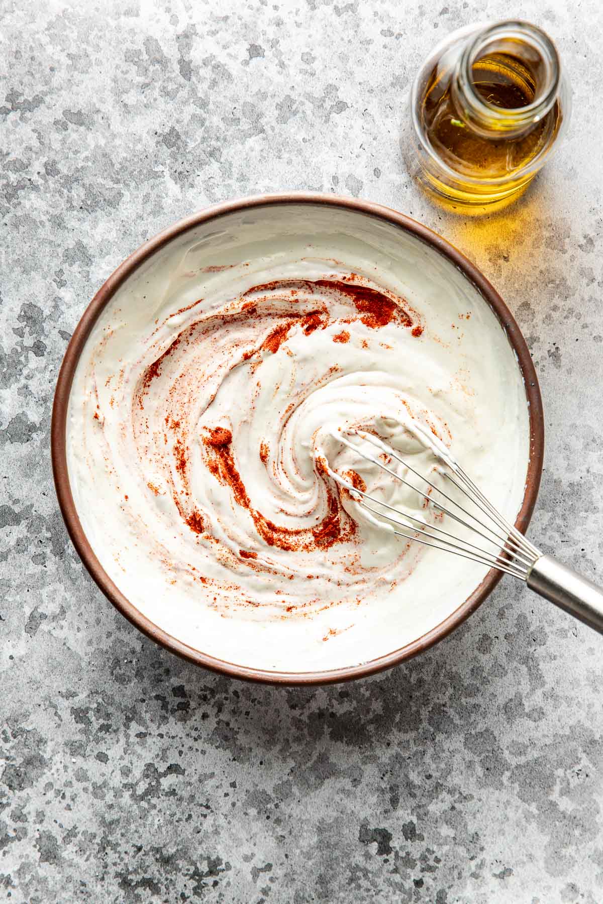 A bowl of creamy white sauce swirled with red spices, being mixed with a whisk; a small bottle of olive oil sits nearby on a textured gray surface.