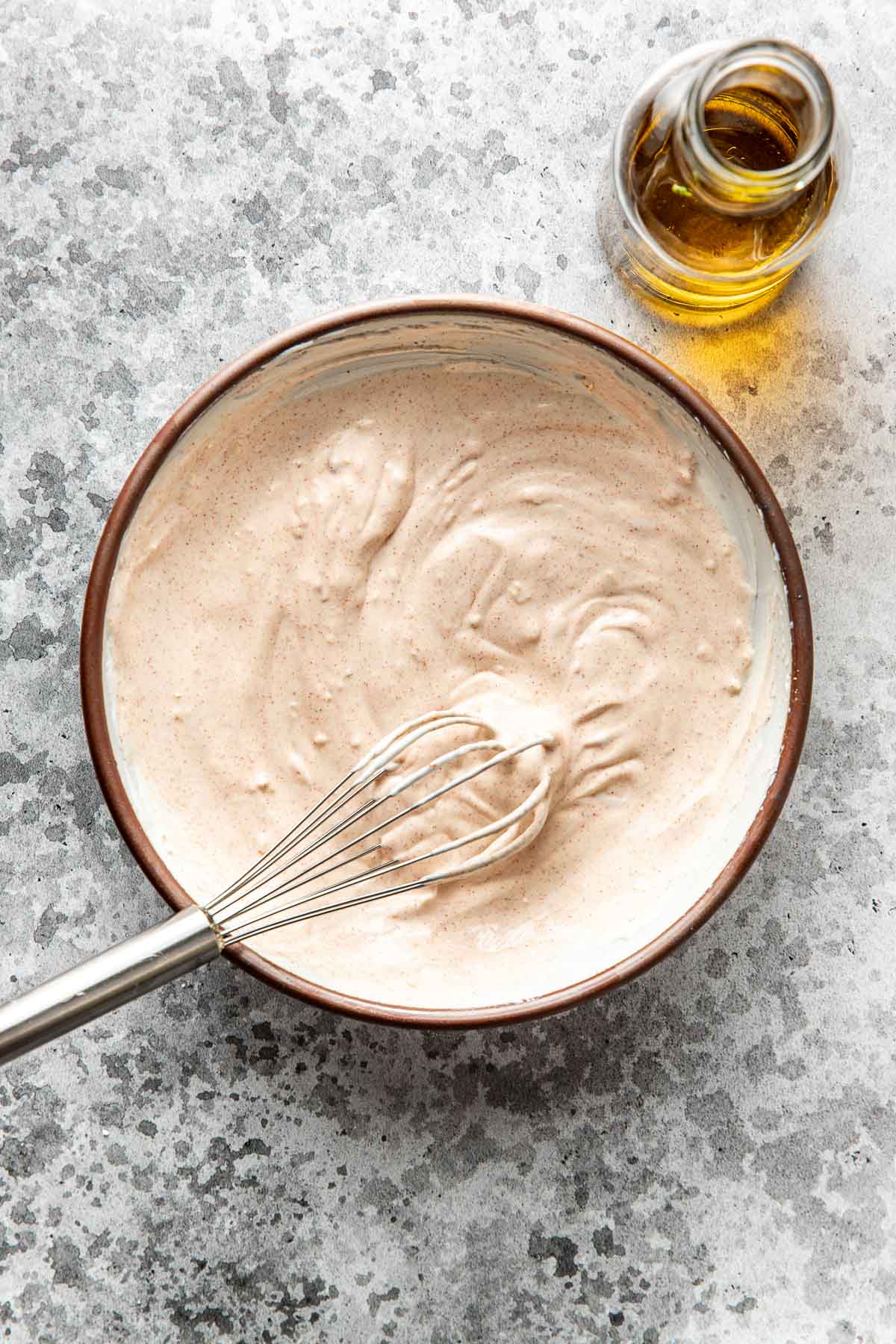 A bowl of creamy sauce being whisked, with a metal whisk resting inside. A small glass bottle of oil sits nearby on a speckled gray surface.
