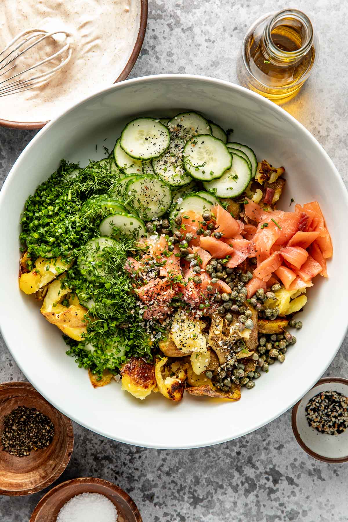 A white bowl filled with sliced cucumbers, fresh herbs, smoked salmon, capers, and roasted potatoes, with cracked pepper on top. Surrounding the bowl are small dishes of black pepper, salt, and a glass bottle of oil.