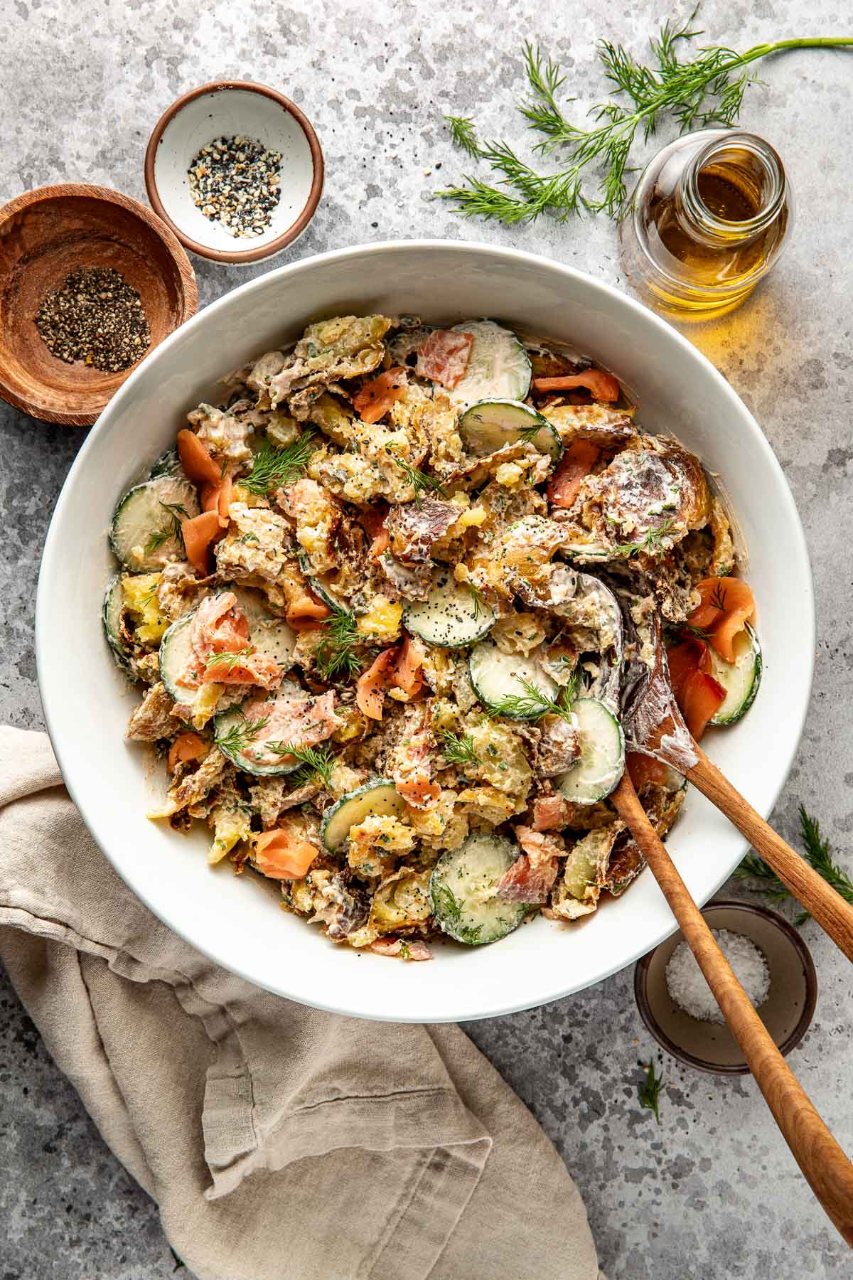 A large white bowl filled with creamy potato salad, sliced cucumbers, smoked salmon, and fresh dill, with wooden serving utensils. Surrounding the bowl are pepper, seasoning, olive oil, and a beige napkin.
