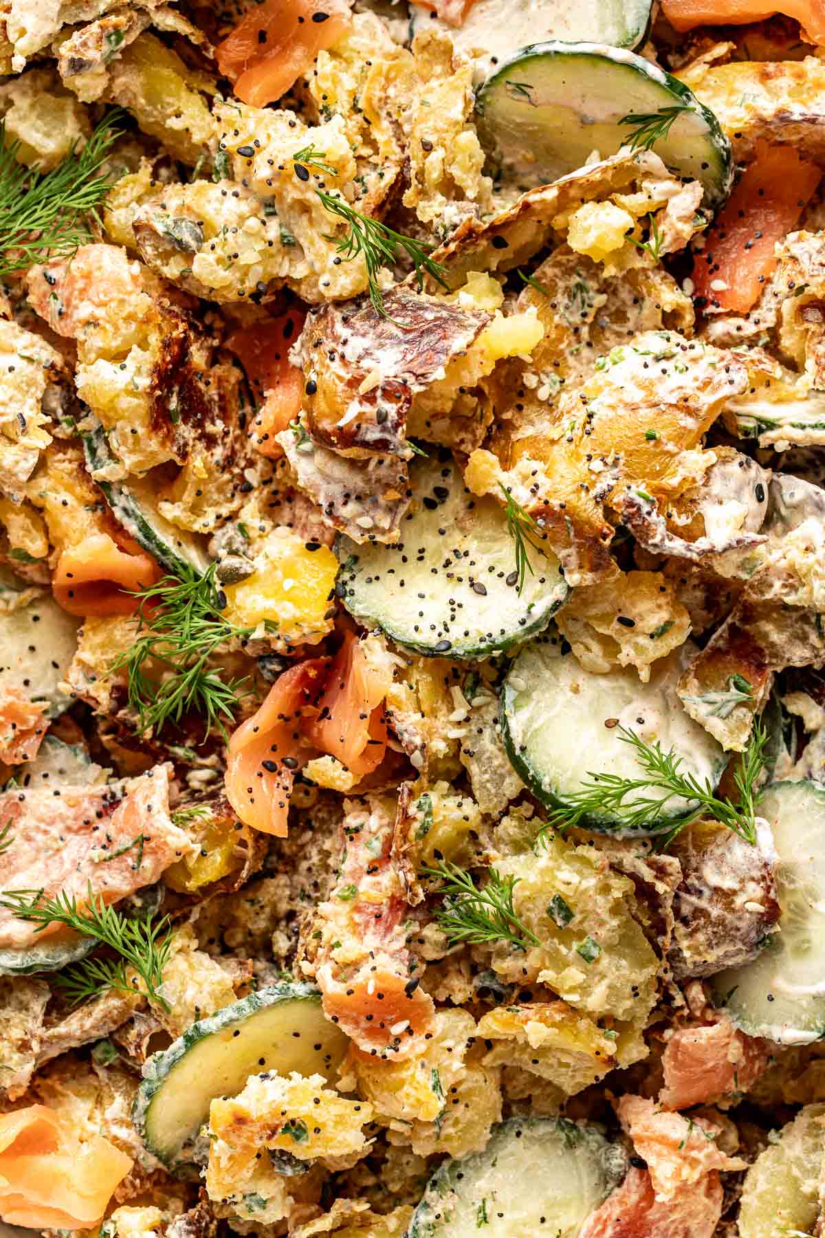 A close-up of a creamy potato salad with sliced cucumbers, dill sprigs, smoked salmon, black pepper, and crispy roasted potato skins. The salad looks fresh and garnished with herbs.