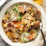 A white plate with a creamy salad containing sliced cucumbers, smoked salmon, fresh dill, and crumbled bagel chips, with a fork on the side. A bottle of olive oil and small bowl of seasoning are nearby.