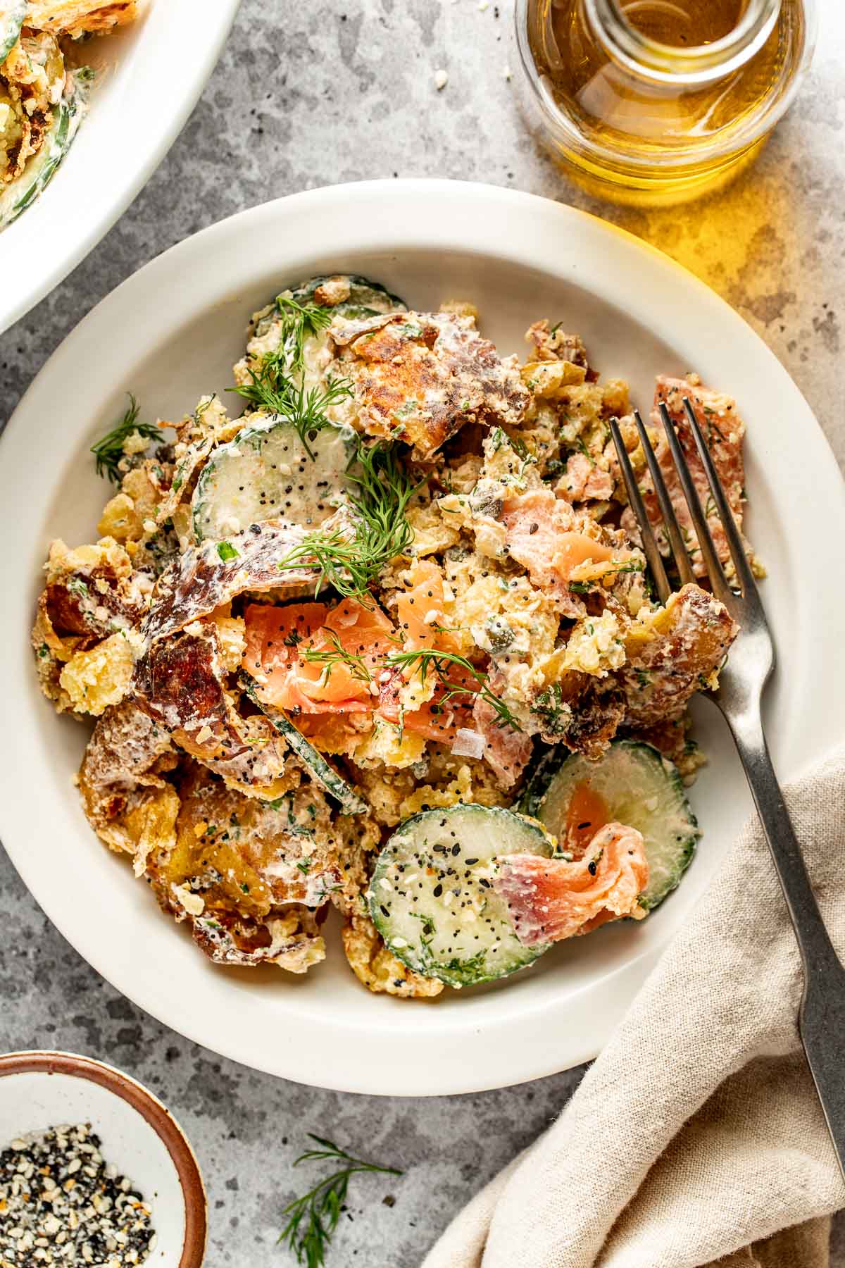 A white plate with a creamy salad containing sliced cucumbers, smoked salmon, fresh dill, and crumbled bagel chips, with a fork on the side. A bottle of olive oil and small bowl of seasoning are nearby.