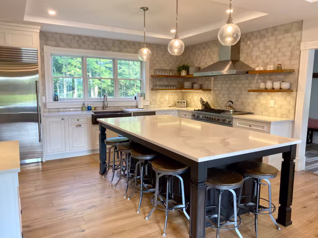 Modern kitchen with a large island, six metal stools, white cabinets, stainless steel appliances, three pendant lights, a tiled backsplash, wood floors, and a window overlooking greenery.